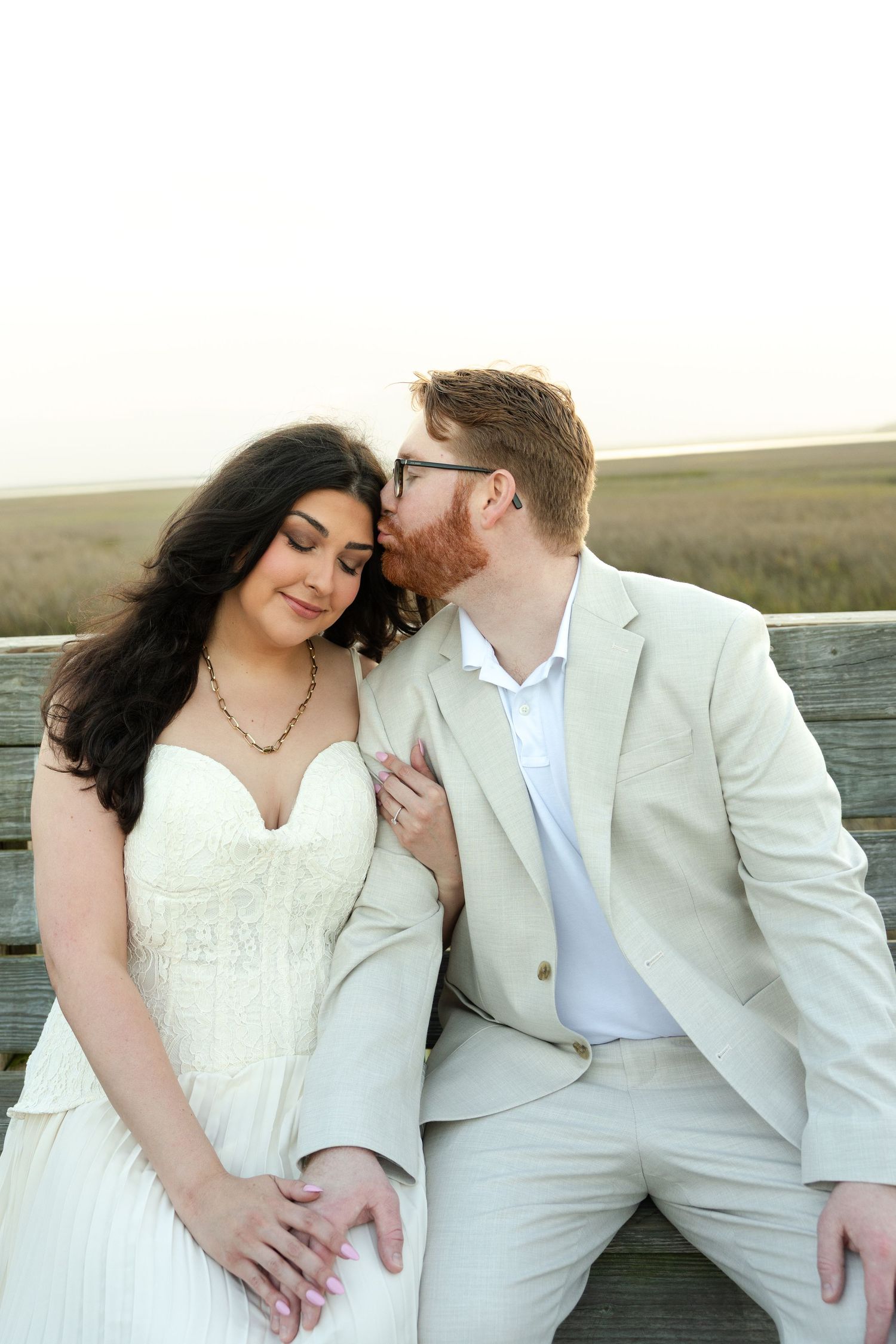 A groom in a beige suit kisses his bride in a white strapless gown on a wooden bench overlooking a marsh.