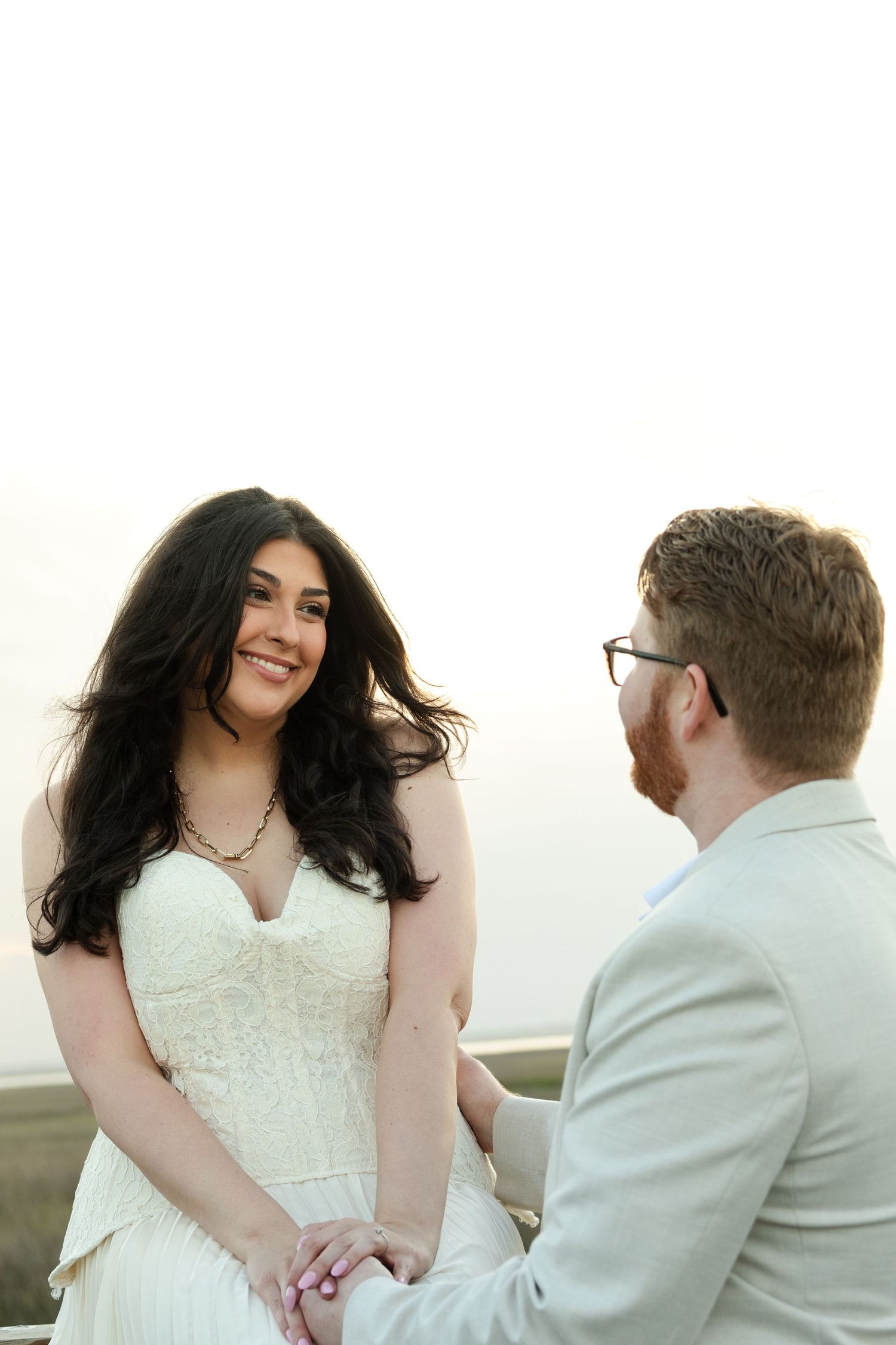 Smiling bride in white strapless gown holds hands with groom in tan suit outdoors during romantic wedding ceremony.