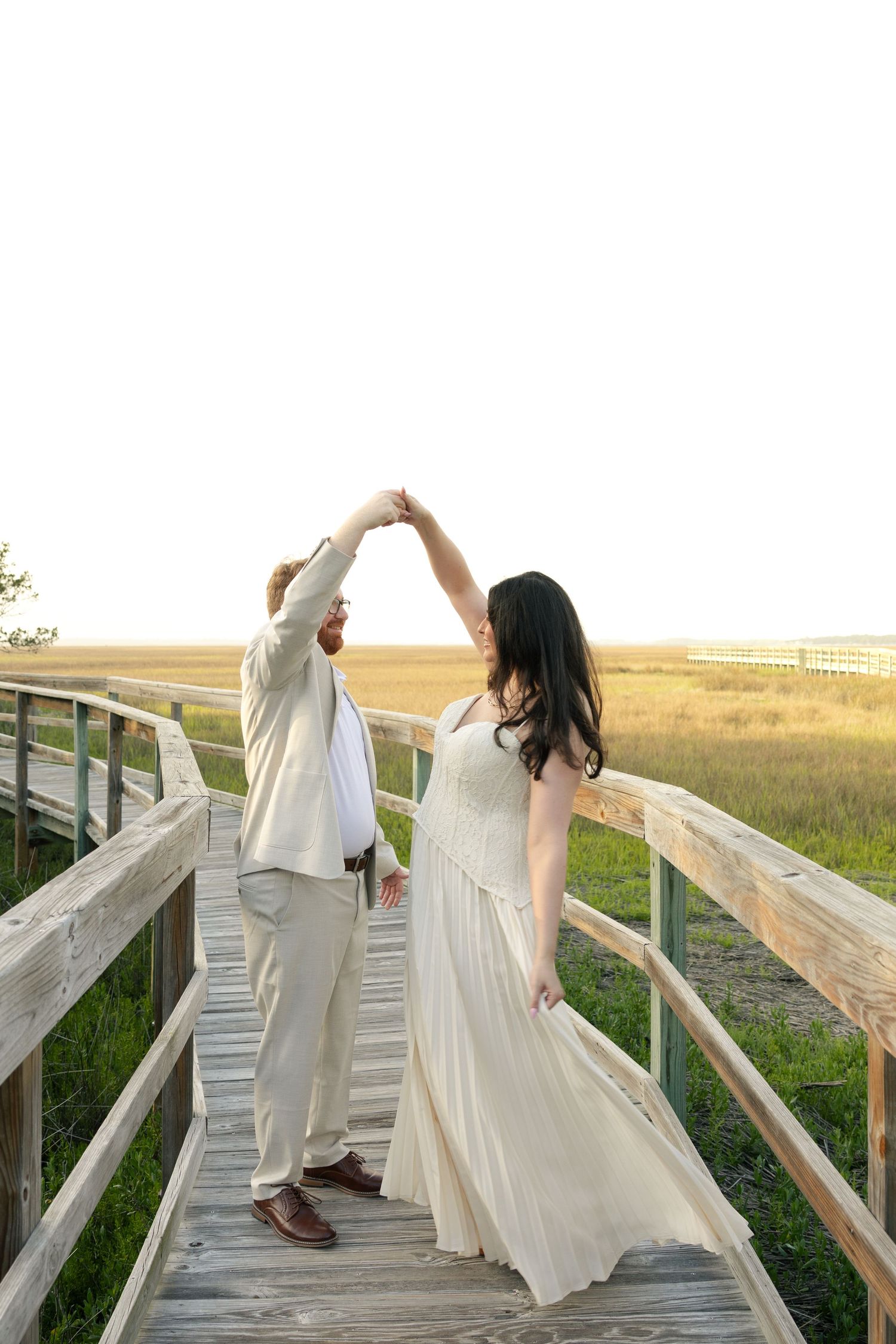 Couple dancing on a wooden boardwalk surrounded by golden marshlands in white and cream outfits at sunset.