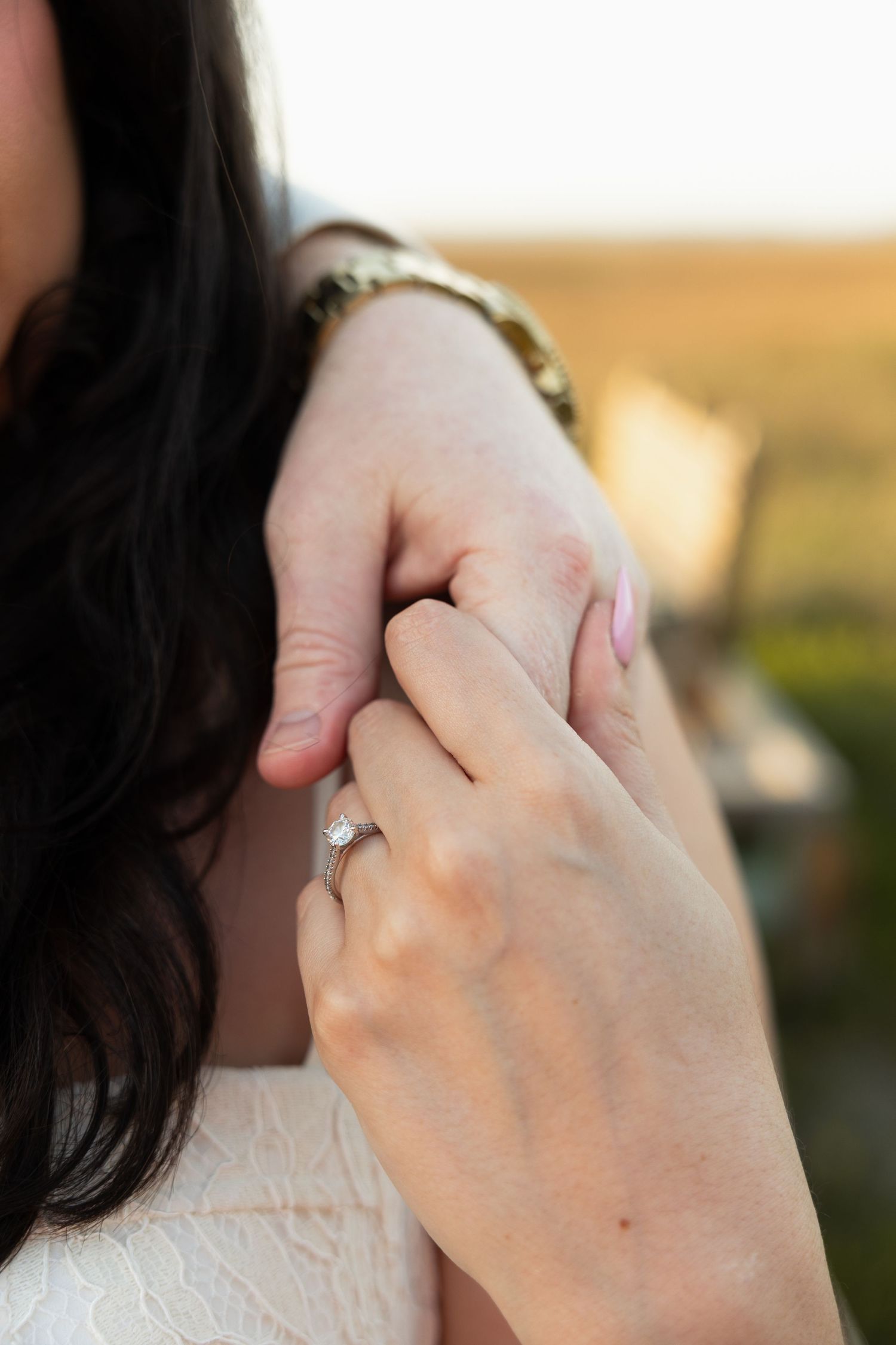 Close-up of two hands holding, featuring a diamond engagement ring, with a gold watch and dark hair visible.