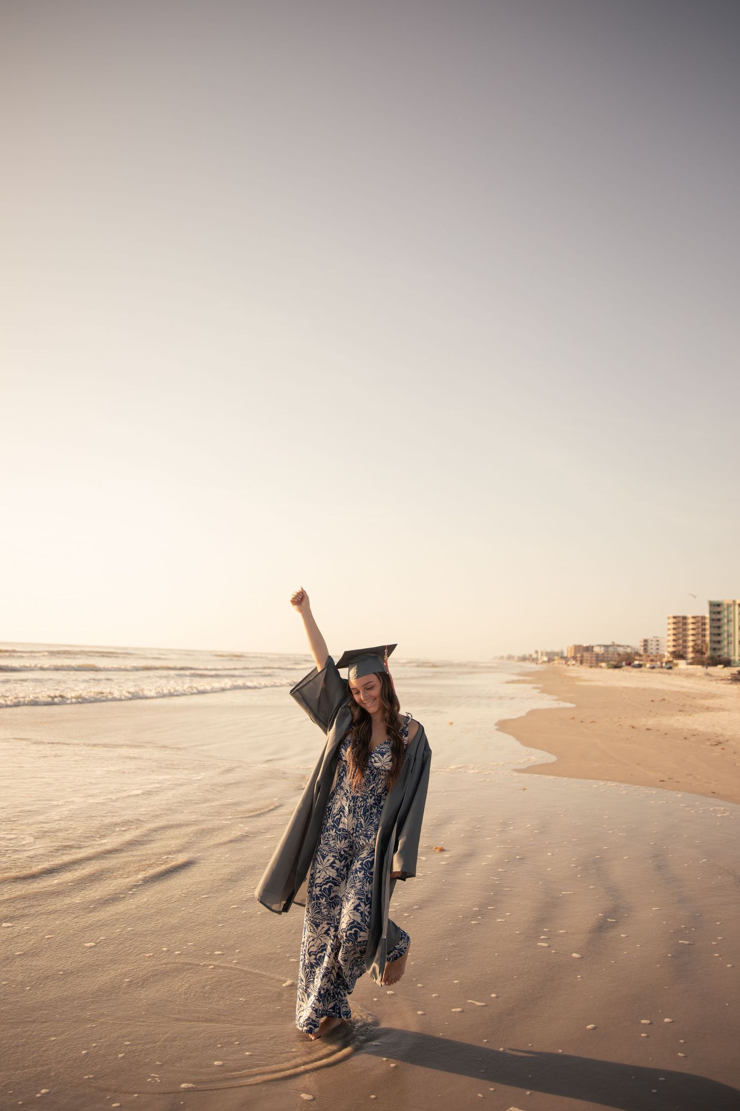 A graduate in cap and gown celebrates on a serene beach at sunset with waves rolling in the background.