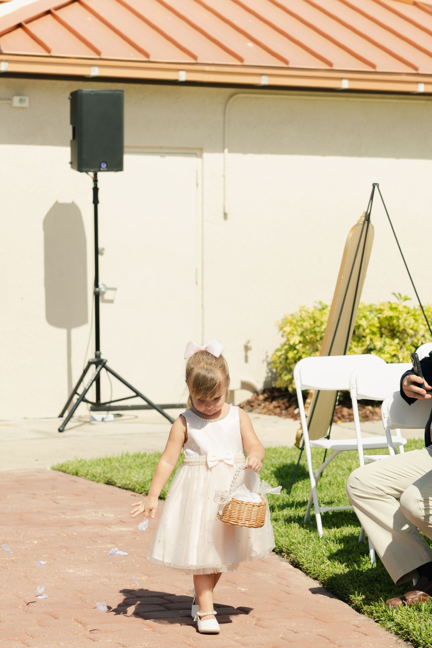 A young flower girl in a white dress scatters petals from a wicker basket along a brick path at an outdoor wedding ceremony.