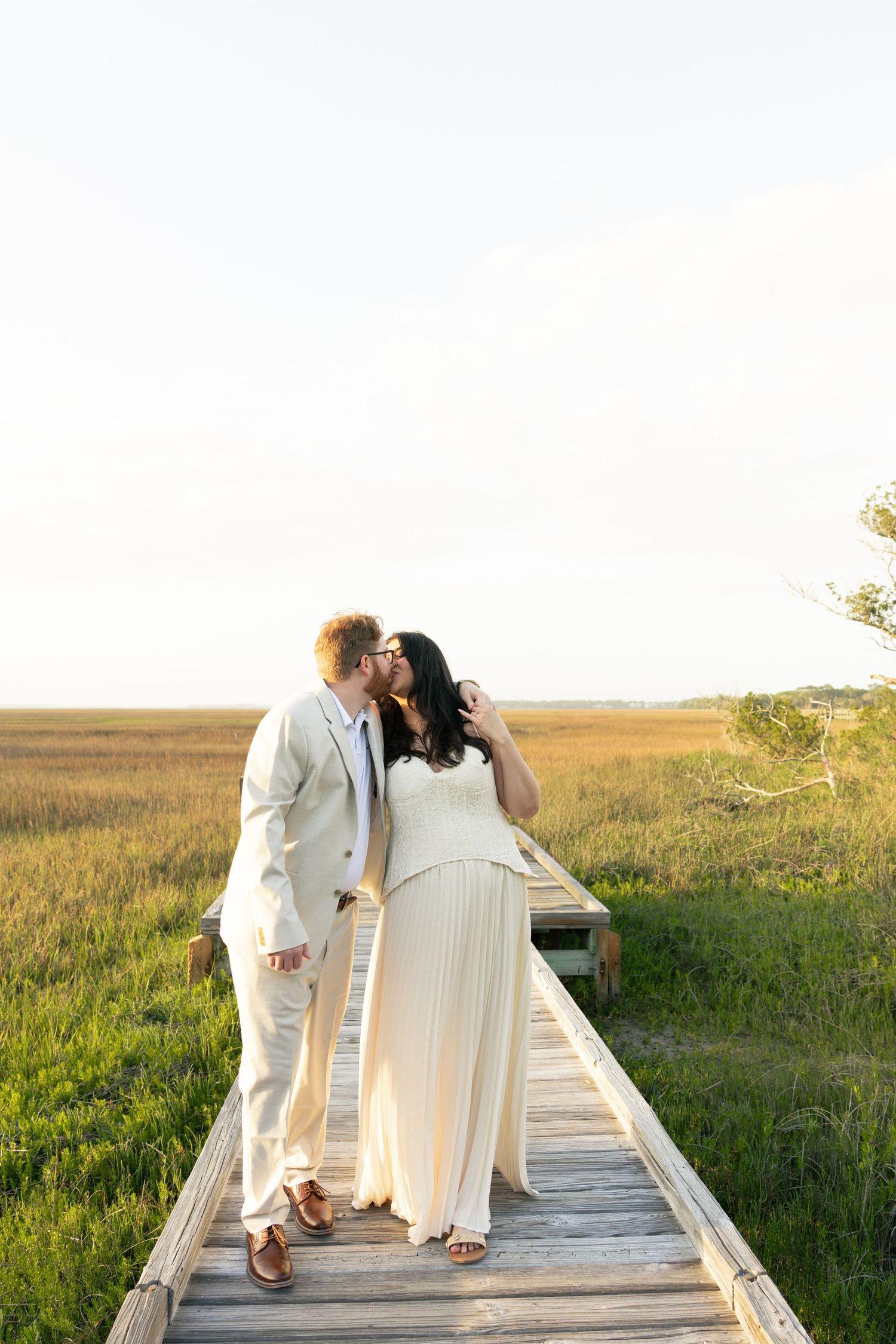Couple sharing a kiss on a wooden boardwalk surrounded by golden marshlands during a romantic outdoor wedding photo session.
