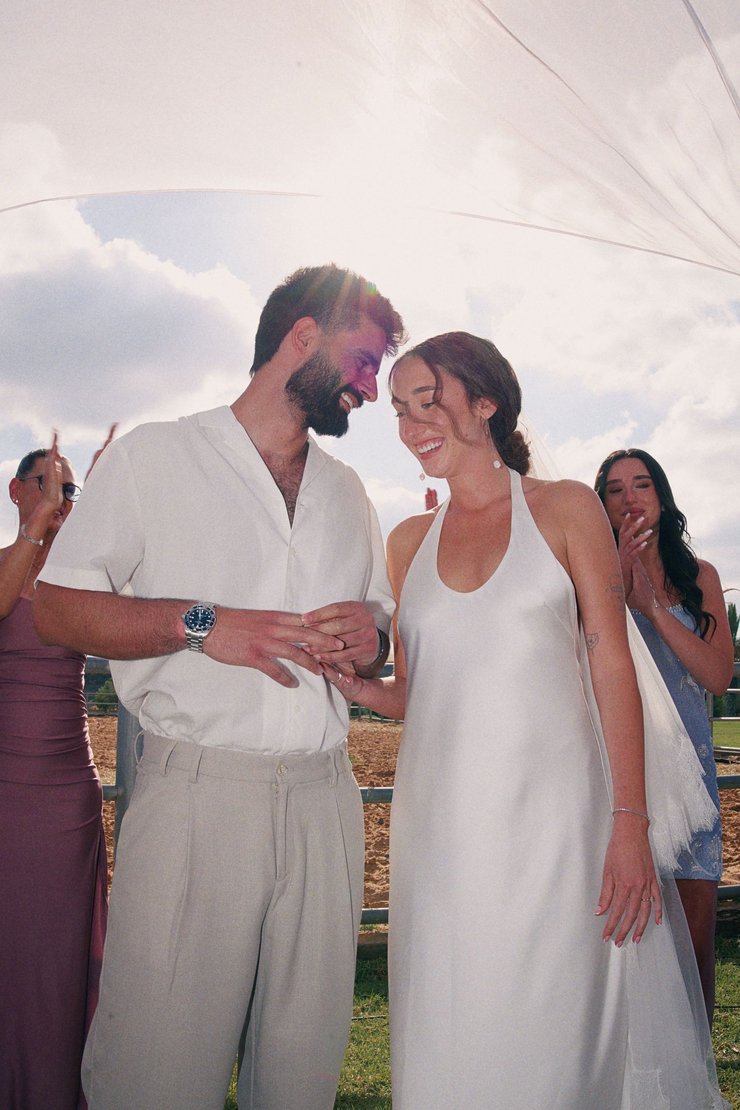Bride and groom exchanging rings outdoors, smiling together at a sunny rustic wedding ceremony.