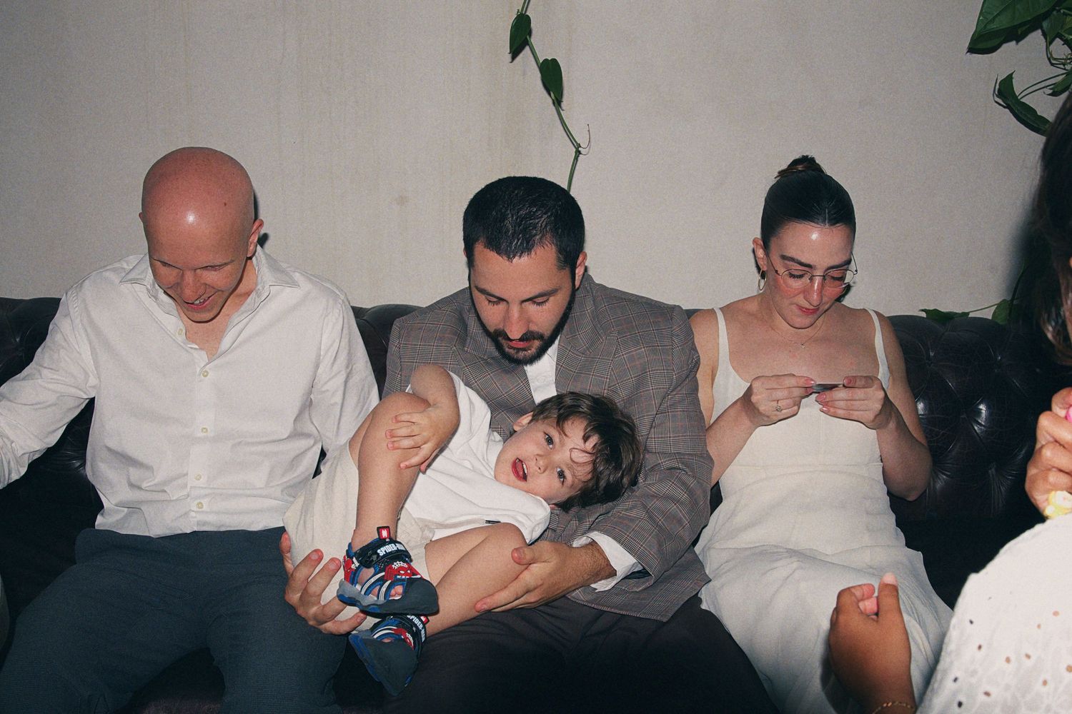 Family sitting on a couch together, man holding a baby, woman in white dress beside him, bald man on the left.
