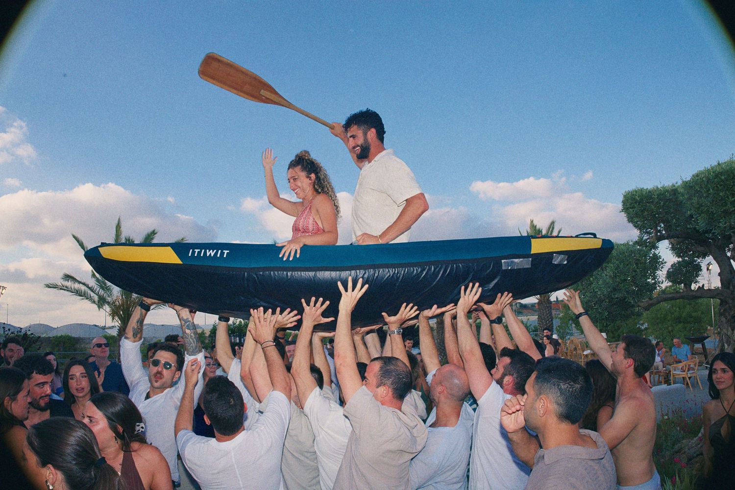 Bride and groom lifted above crowd in a canoe during festive outdoor wedding reception.