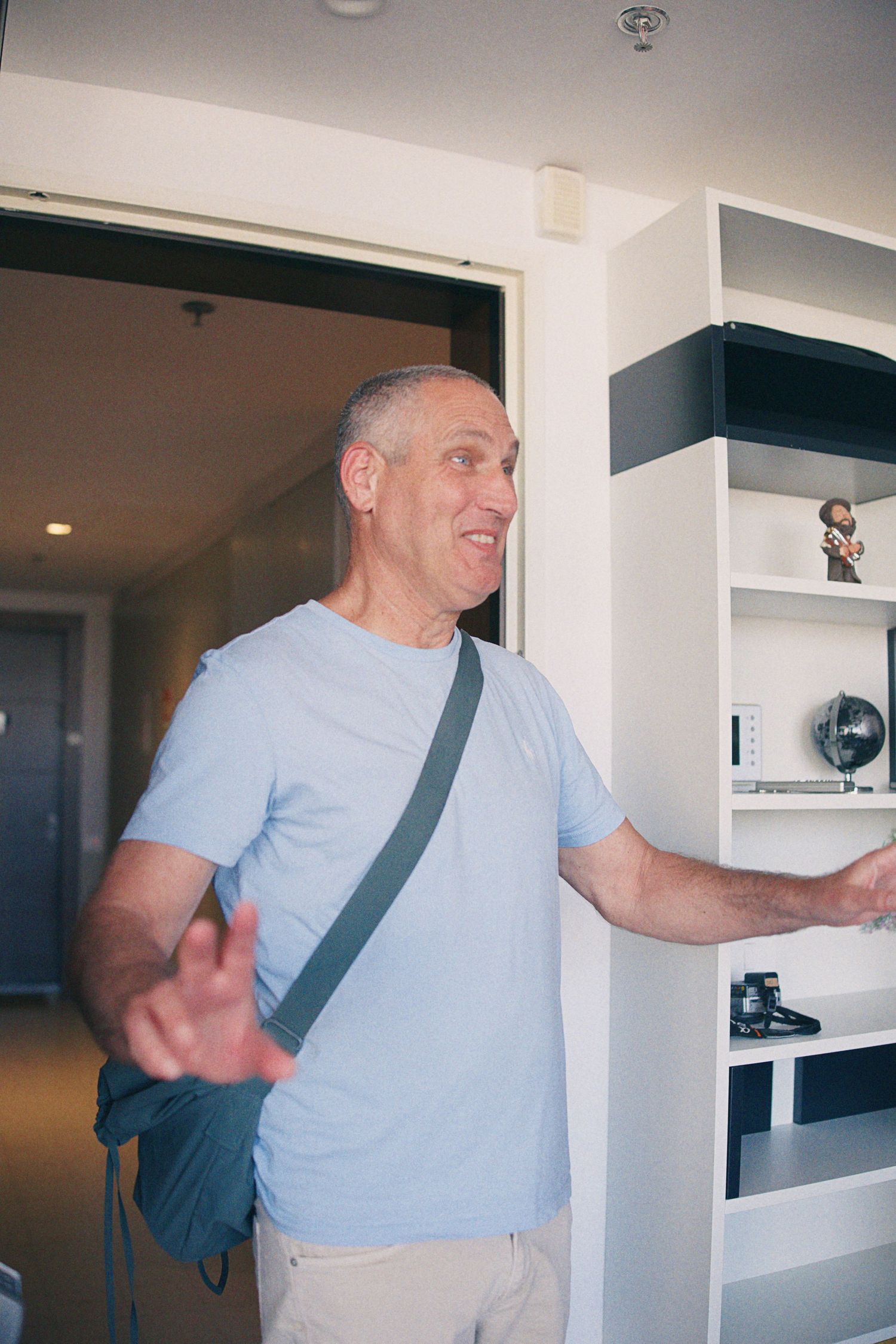 Middle-aged man in blue t-shirt with crossbody bag gestures near white bookshelf indoors.
