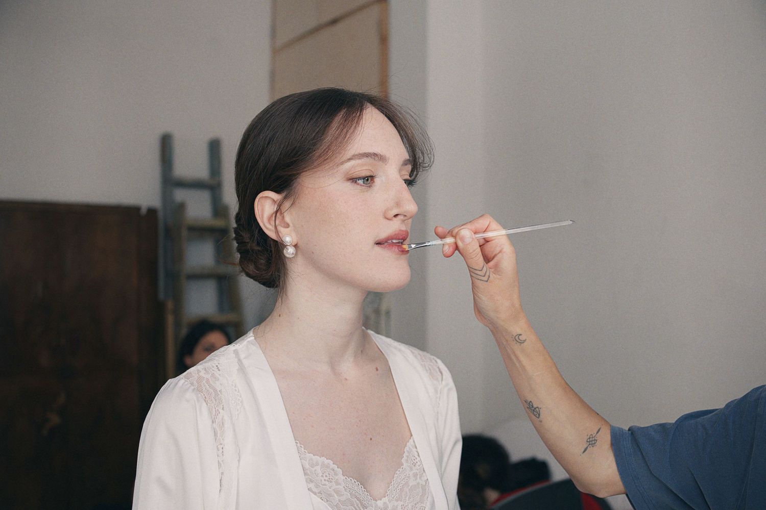 A makeup artist applying lipstick to a young woman in a white robe during bridal preparation.
