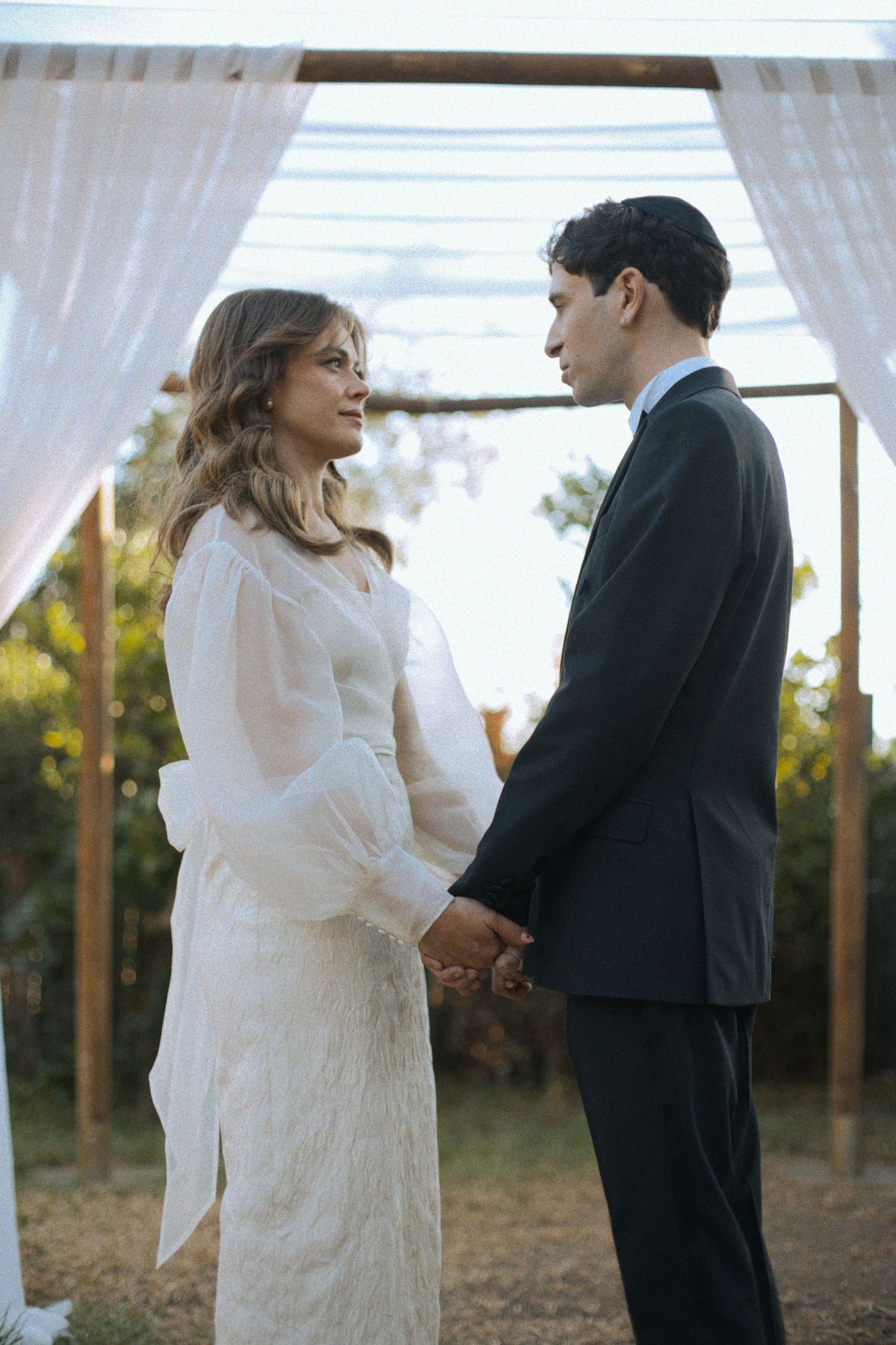 Bride in white lace dress and groom in black suit standing under white draped ceremony arch.