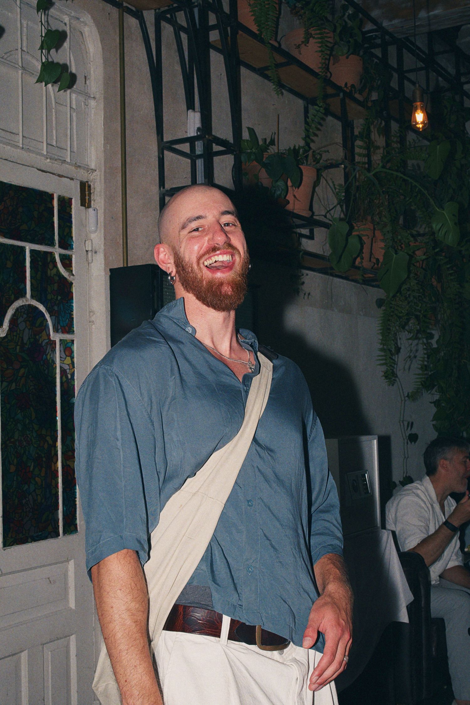 Smiling bearded man in blue shirt with crossbody bag laughing in a plant-decorated restaurant.