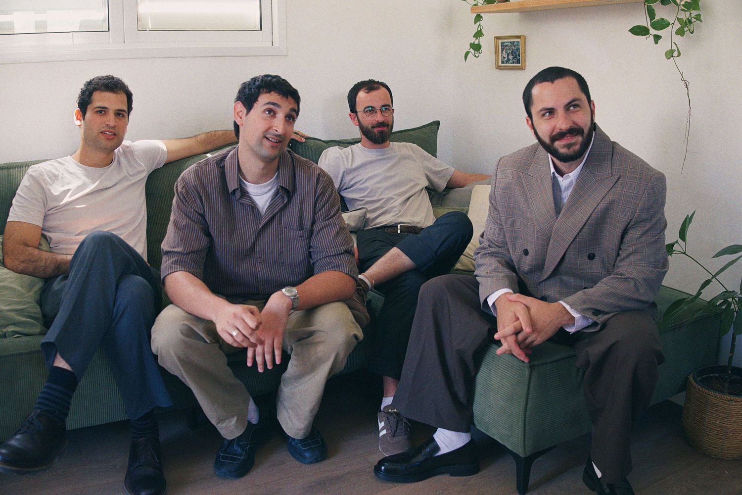 Four men sitting together on a green sofa in a bright modern room, dressed in casual and smart-casual attire.