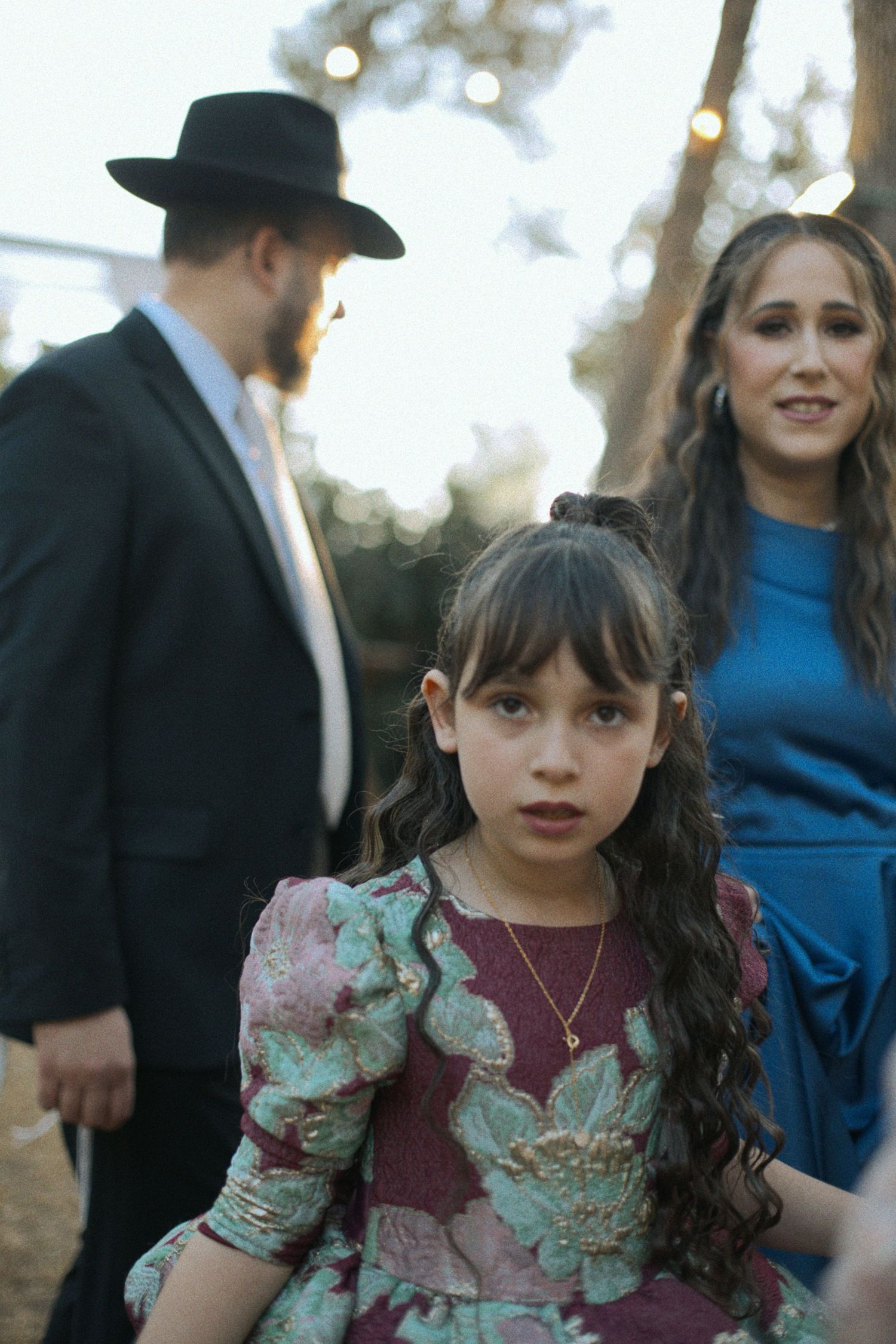 Young attendee in floral dress at outdoor wedding celebration with guests mingling in background.