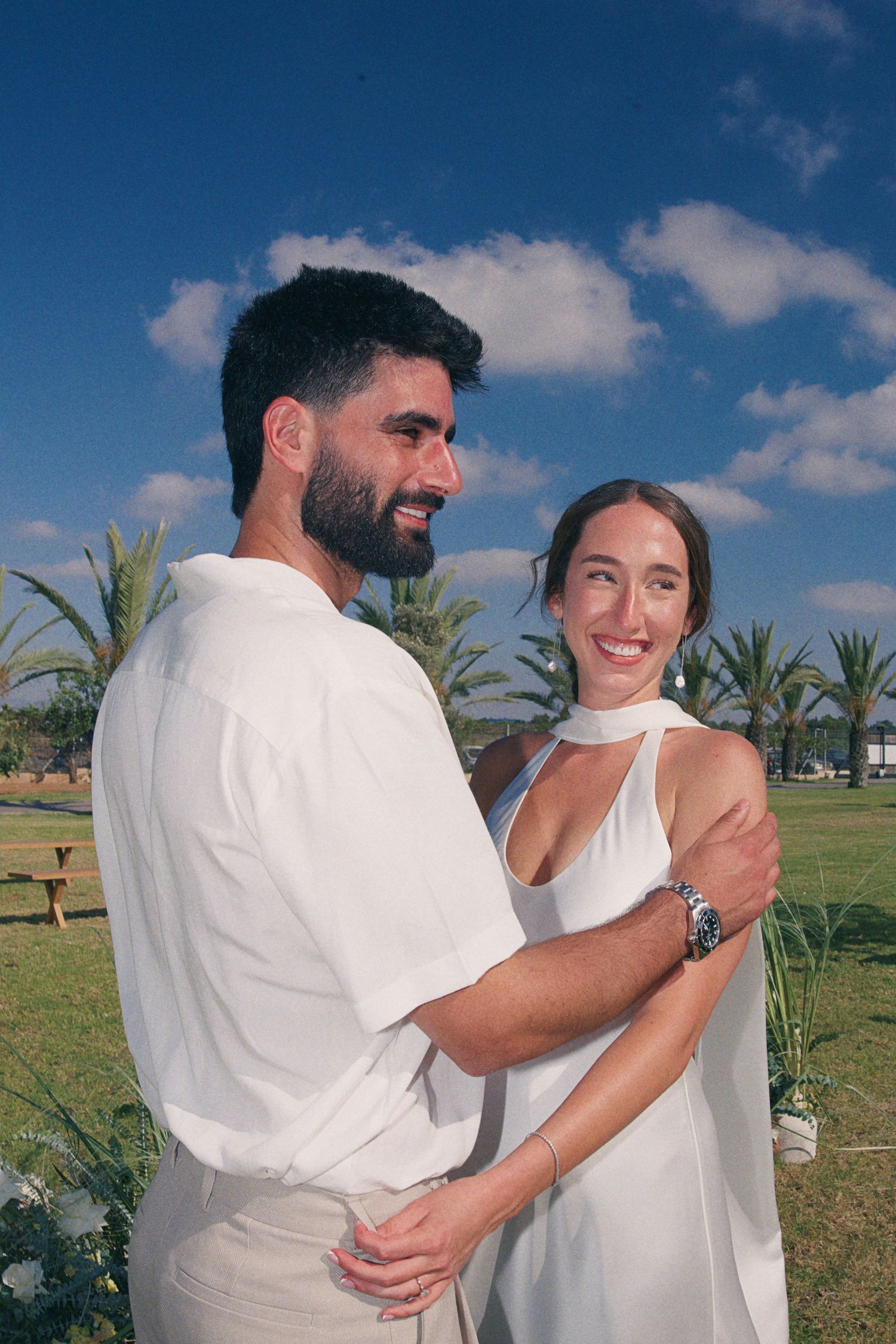 Couple in matching white outfits embrace and smile outdoors with palm trees and blue sky behind them.