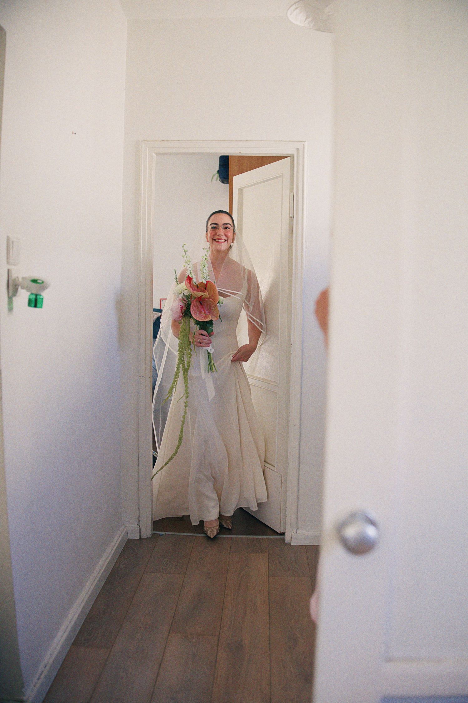 Smiling bride in white gown and veil holding a pink floral bouquet standing in a doorway.