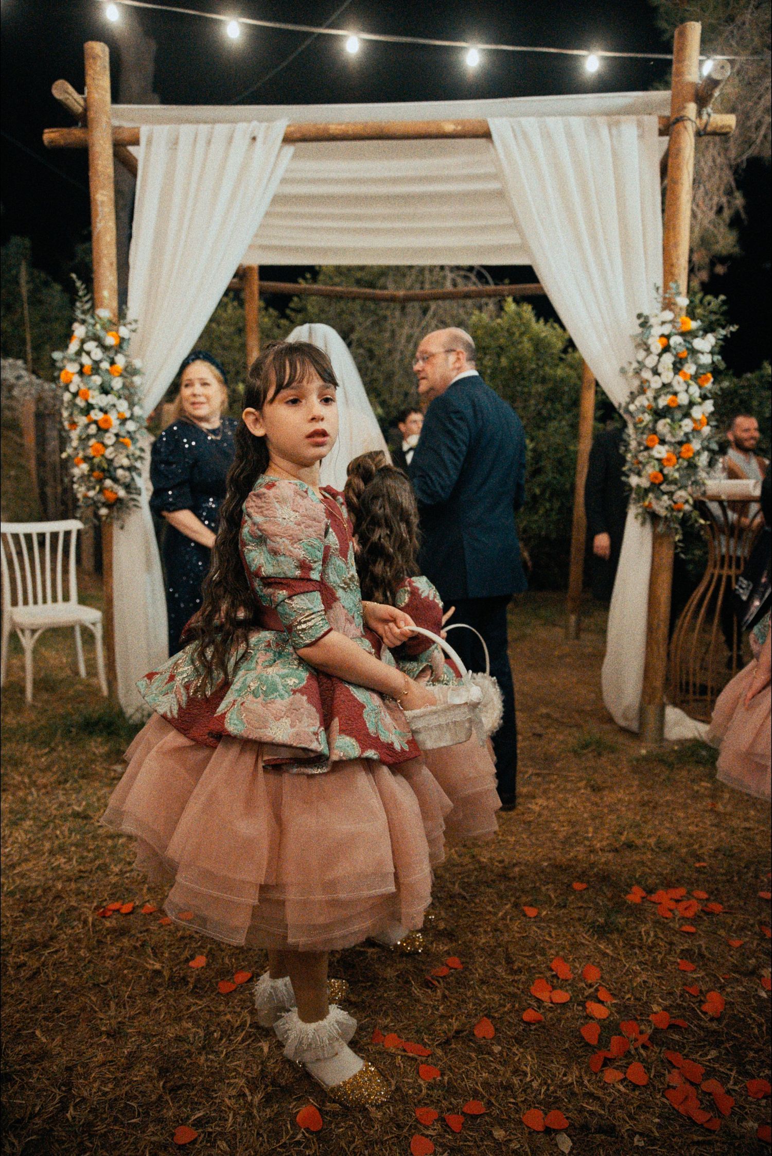 Young flower girl in a tulle dress twirls at an outdoor wedding ceremony under a wooden arch with white draping.