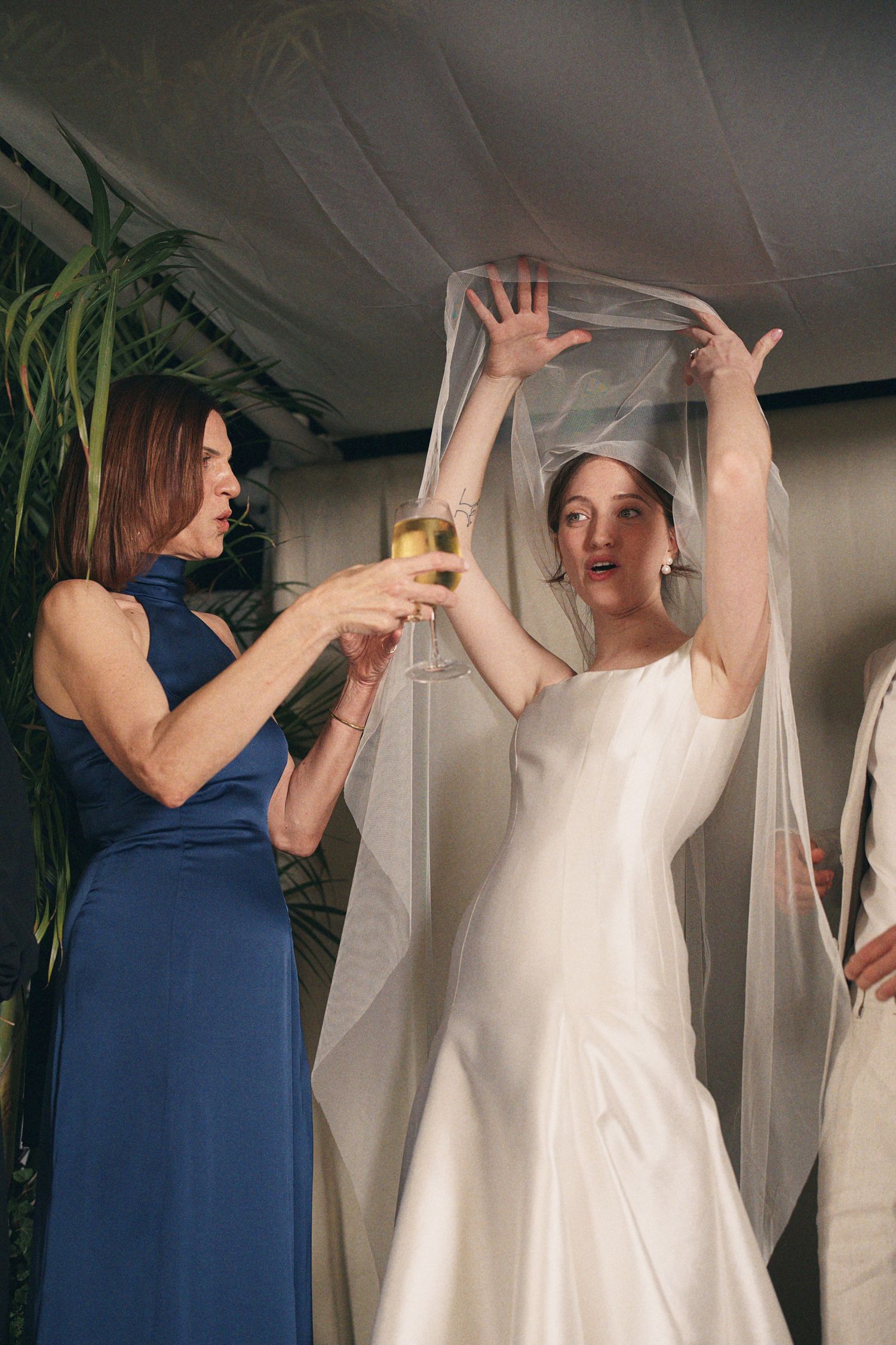 Bride in white gown lifting veil with bridesmaid in blue dress holding champagne glass at wedding celebration.