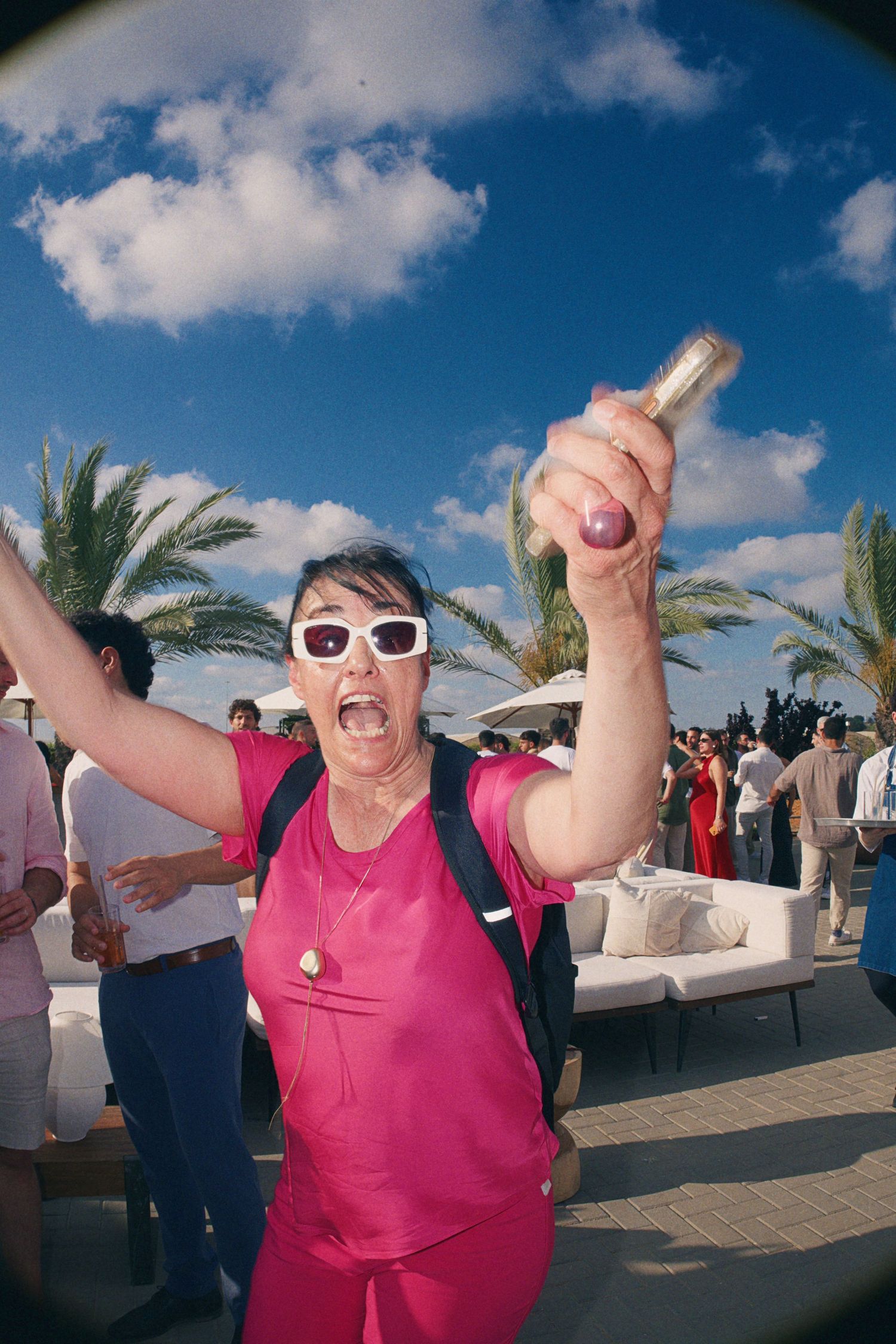 Woman in pink outfit raises bottle celebrating outdoors at tropical venue with palm trees and blue sky.
