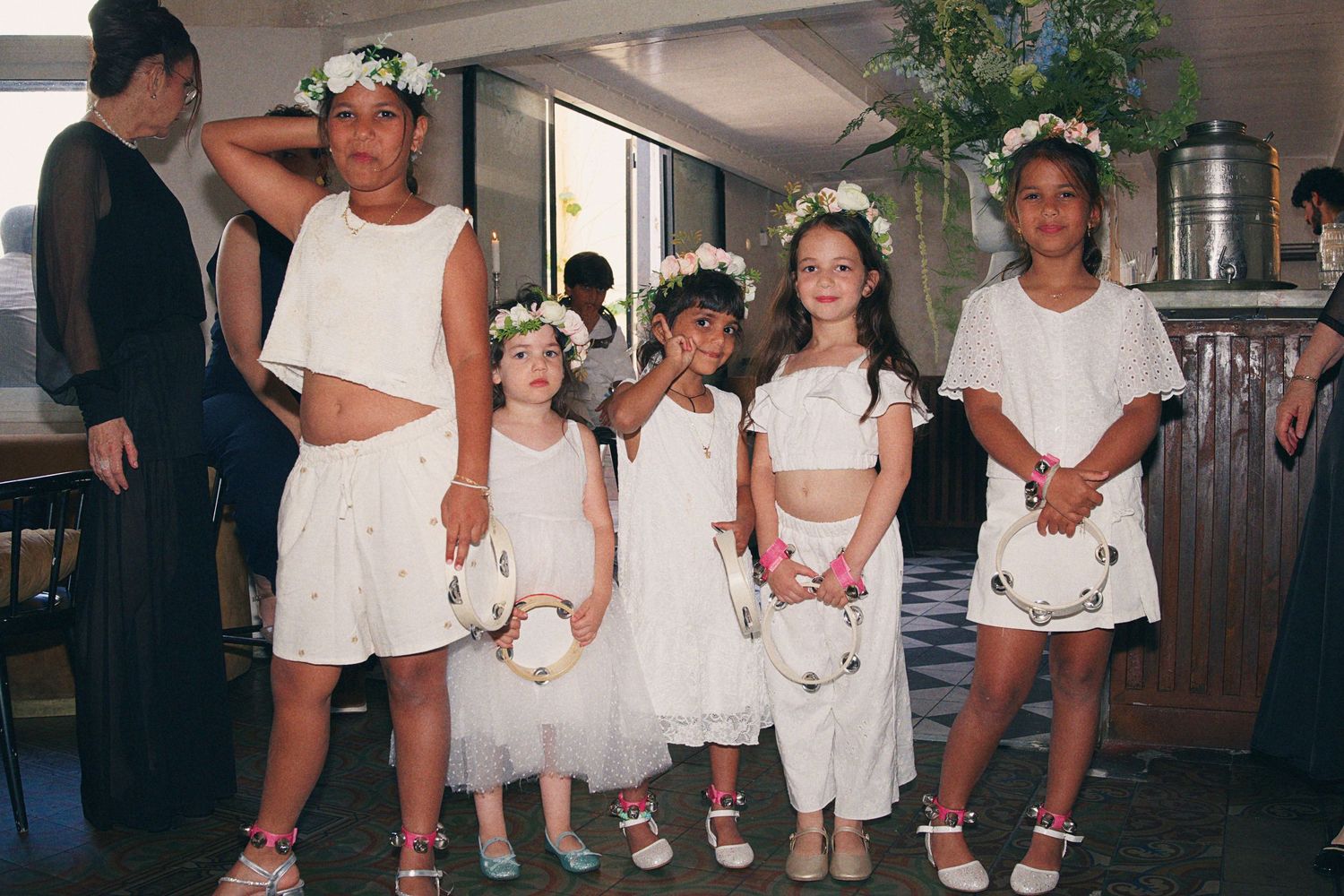 Five children in matching white outfits and floral crowns pose together at a wedding ceremony.