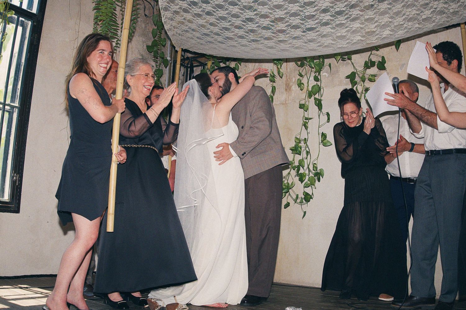 Bride and groom celebrate under a floral chuppah surrounded by clapping guests at a Jewish wedding ceremony.