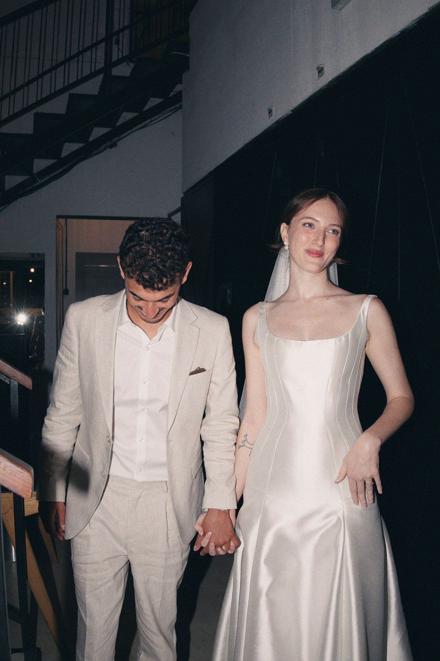 Bride in silver satin gown and groom in linen suit holding hands walking down stairs at night.