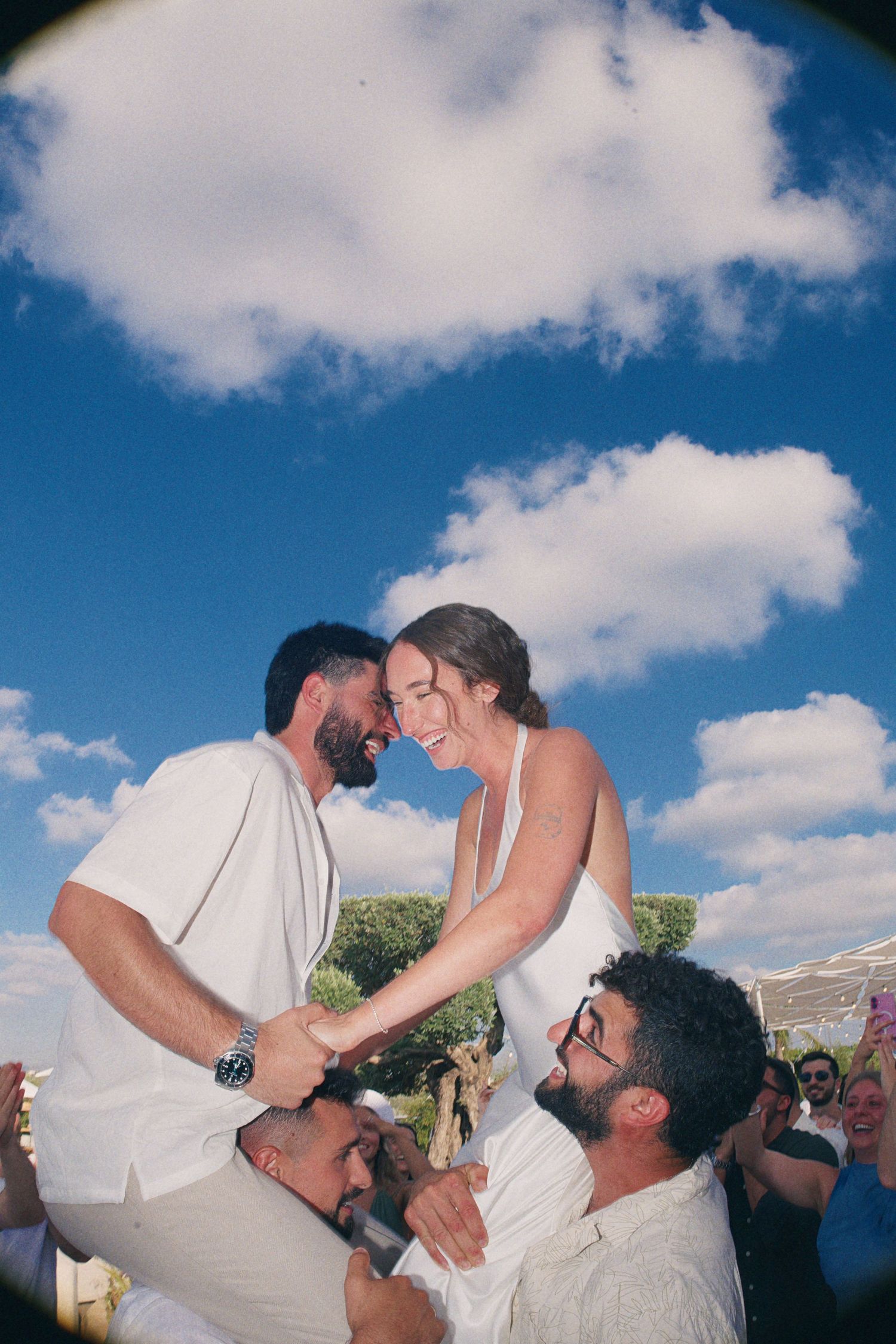 Joyful couple in white embraces outdoors under blue sky at festive celebration event.