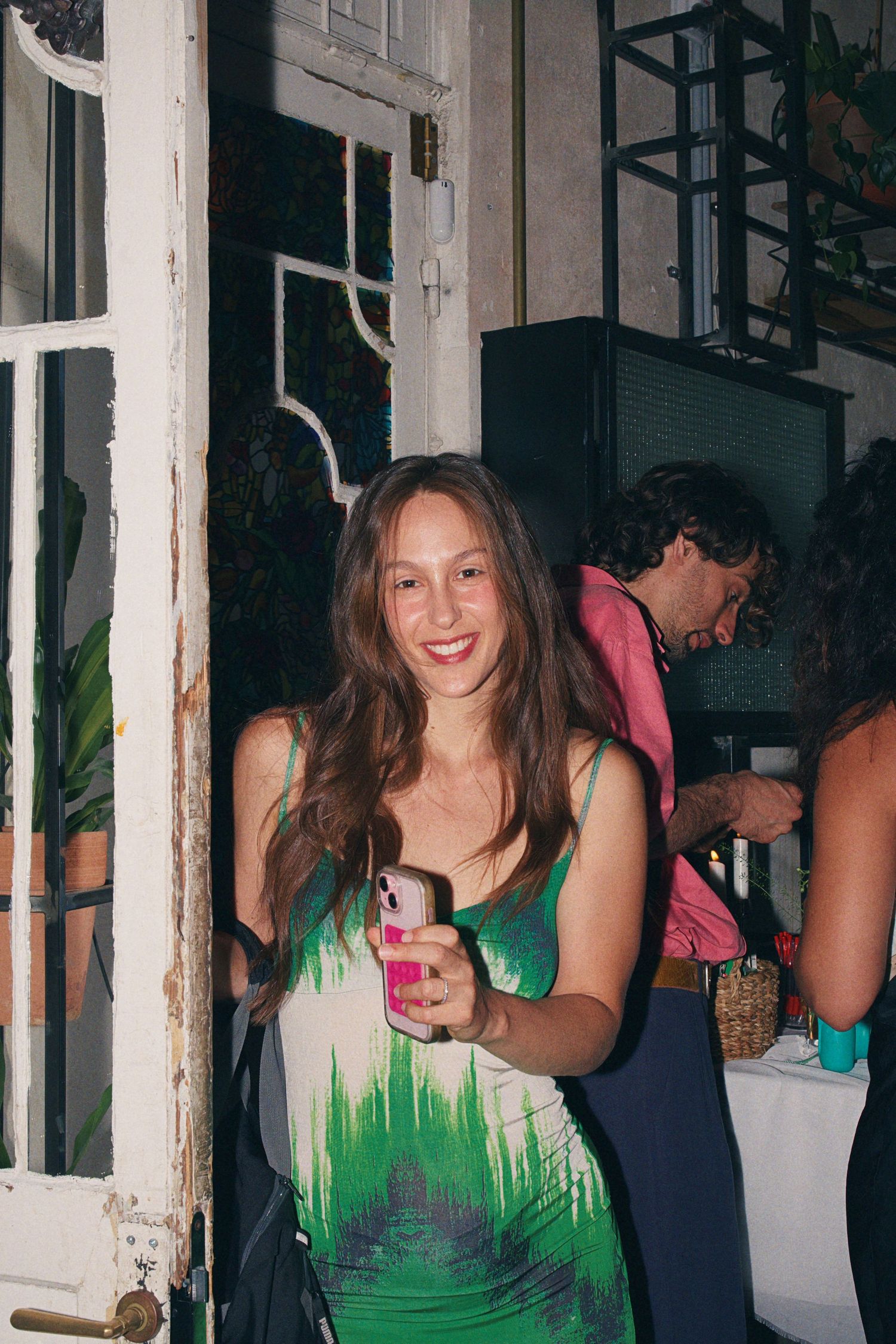 Smiling young woman in green tie-dye dress holding pink phone case at indoor social gathering.