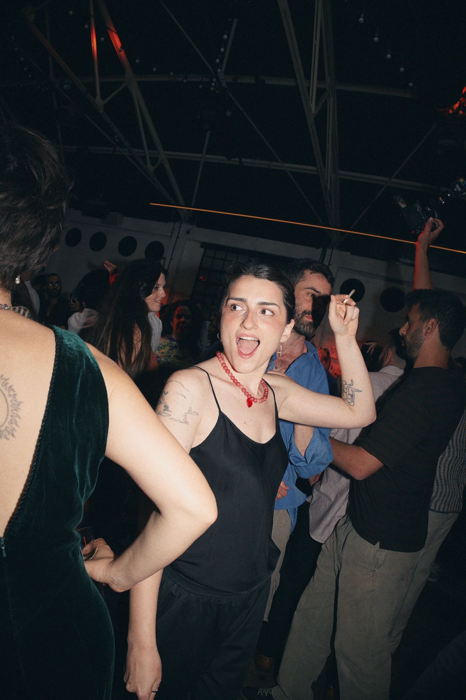 Young woman dancing at a nightclub party, wearing a black dress with tattoos and a red necklace, surrounded by crowd.