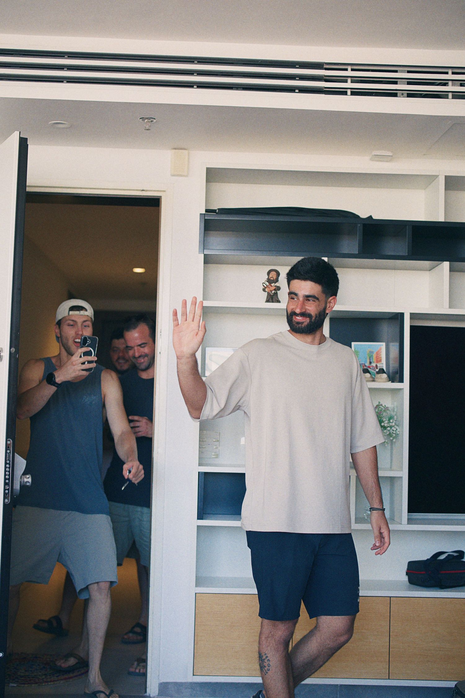 Bearded groom in beige oversized t-shirt waves while friends photograph him in a modern hotel room.