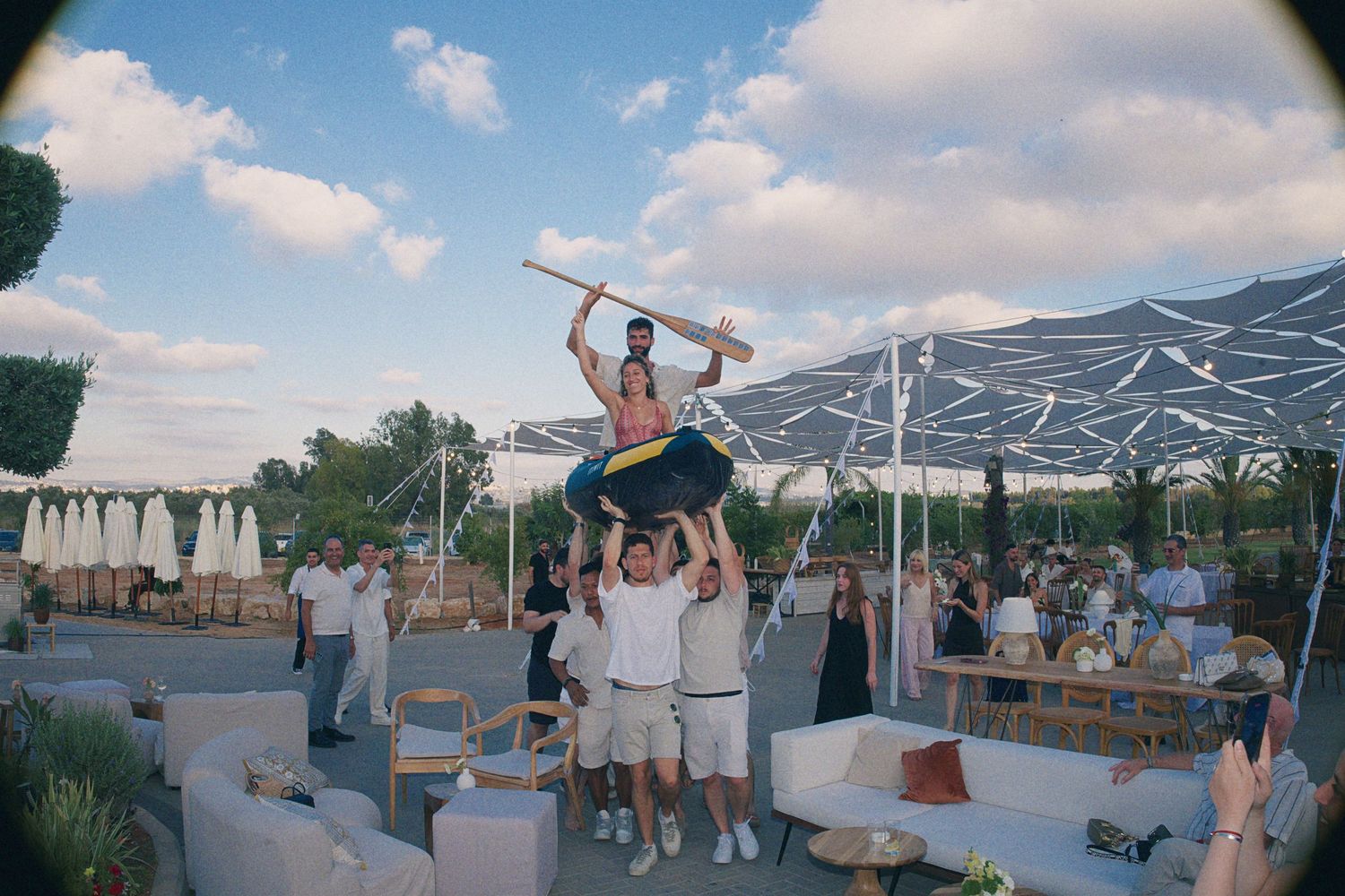 Man lifted high by friends at outdoor wedding venue with white umbrellas and lounge seating visible.