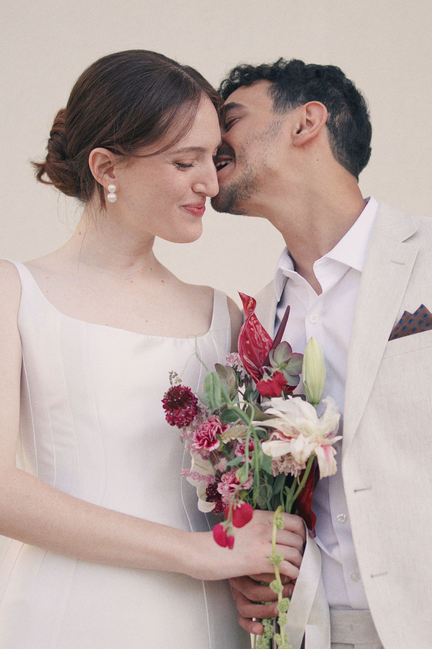 A groom in a beige suit kisses his bride wearing a white dress while holding a red and pink floral bouquet.