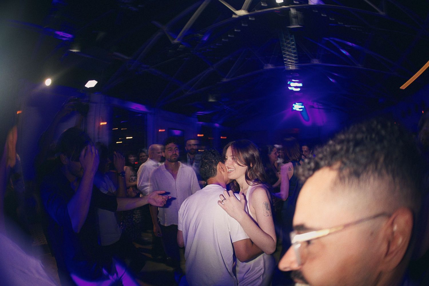 Couple sharing a romantic kiss on a crowded nightclub dance floor illuminated by purple and blue lights.