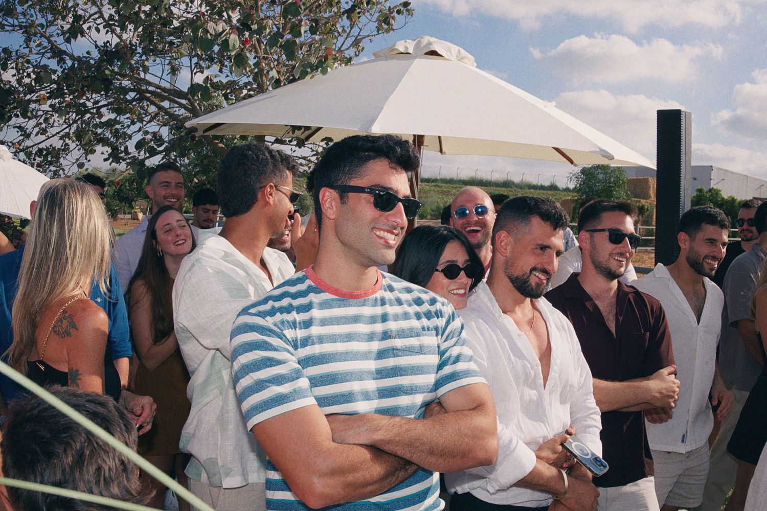 Smiling man in striped shirt with crossed arms at an outdoor social gathering on a sunny day.