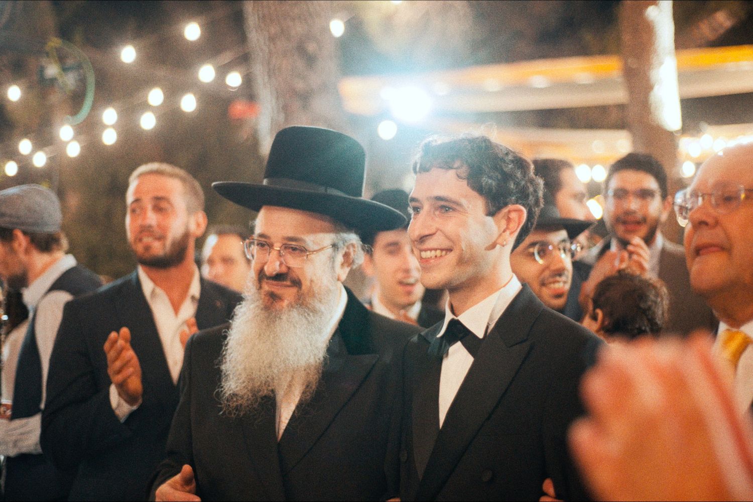 A crowd of guests in dark formal attire and hats celebrate at an outdoor evening event.
