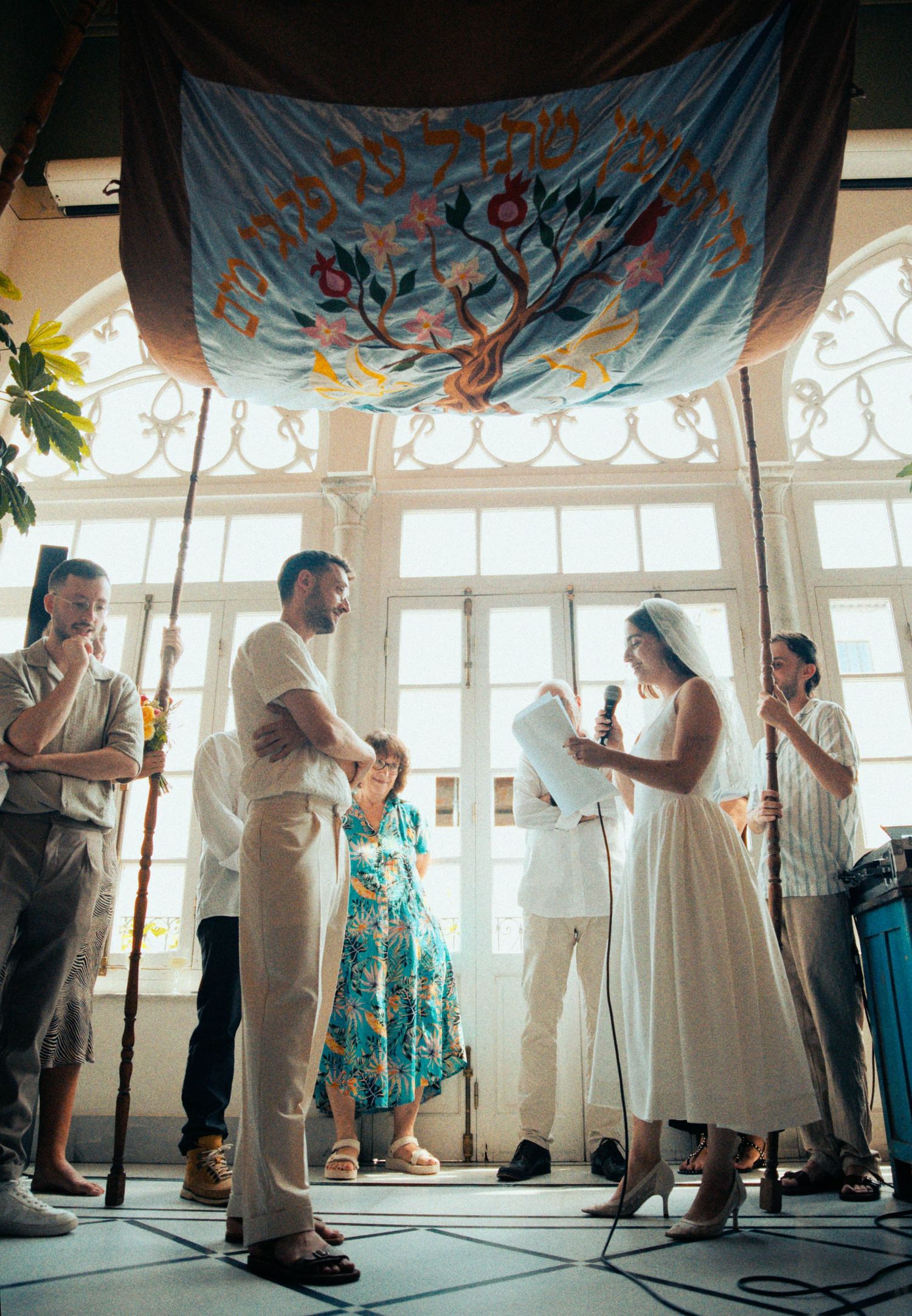 Wedding celebration with guests dancing under decorative banners in a sunlit room.