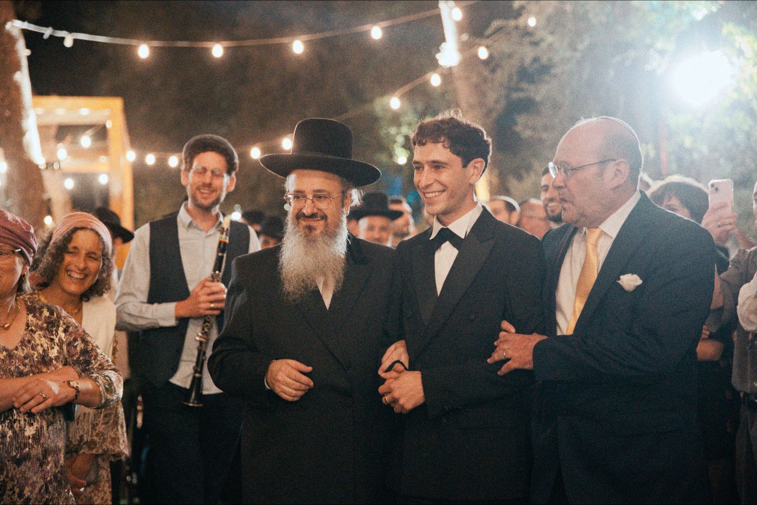 Festive outdoor wedding reception with string lights and dancing guests in formal attire.