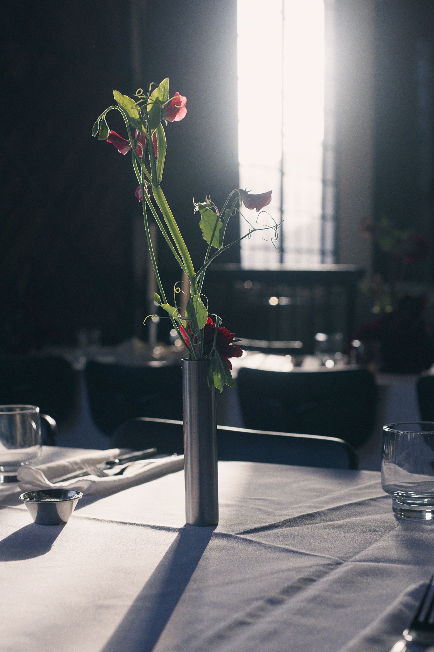 Elegant restaurant table setting with silver vase holding red sweet peas, glassware, and white tablecloth in moody lighting.