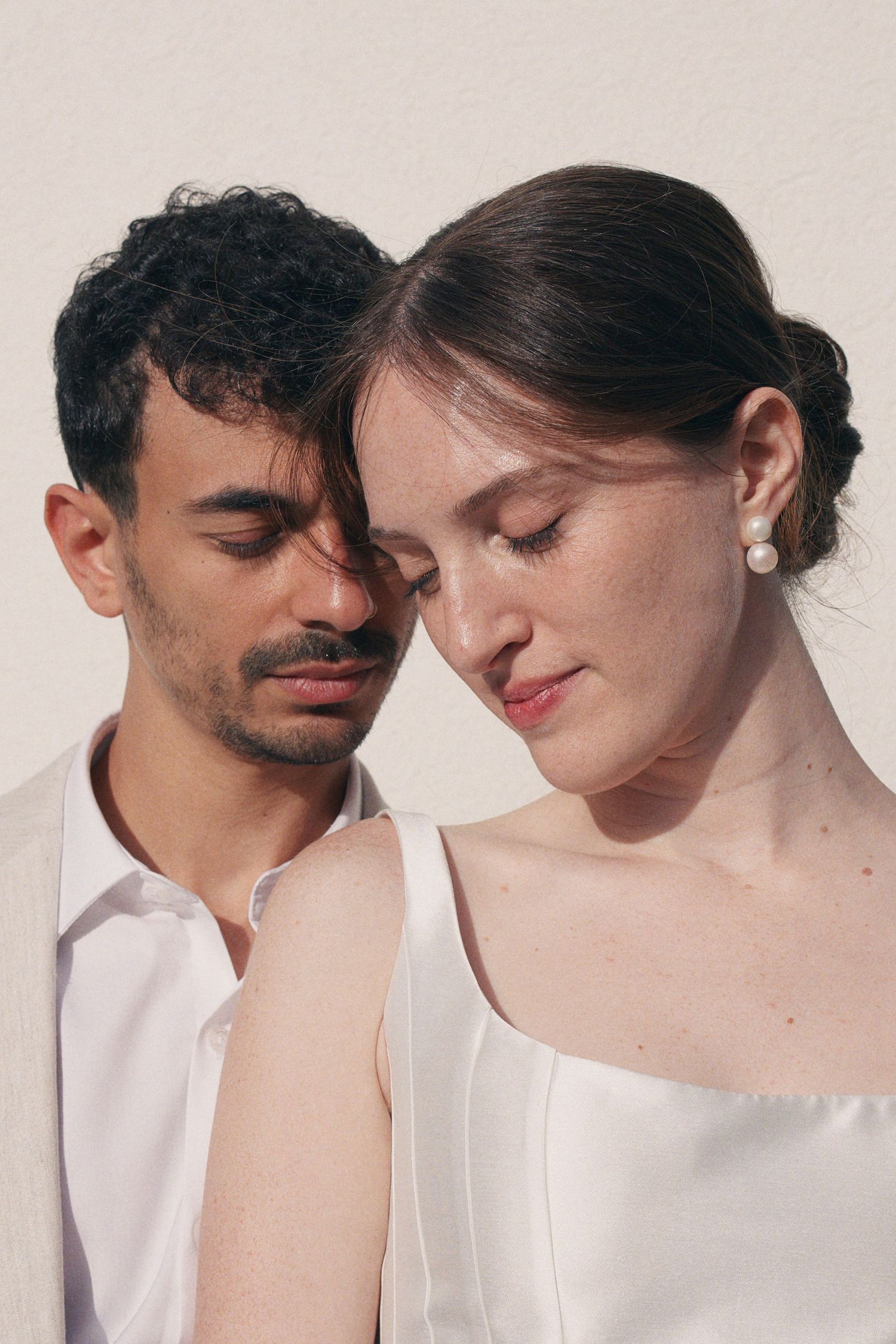 Intimate close-up of wedding couple in white attire with eyes closed, touching foreheads tenderly.