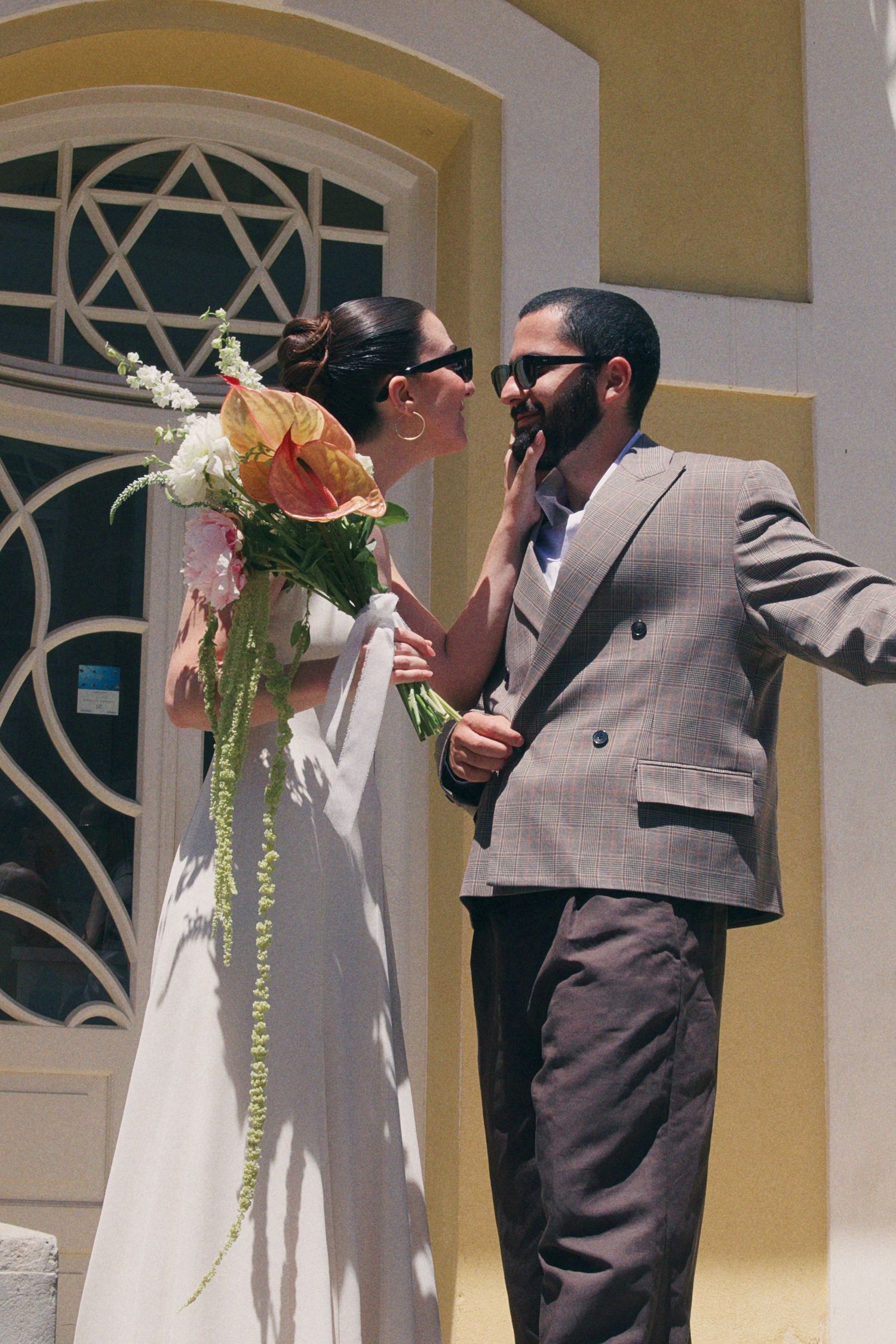 Bride in white dress holding floral bouquet touches grooms face outside sunny building.