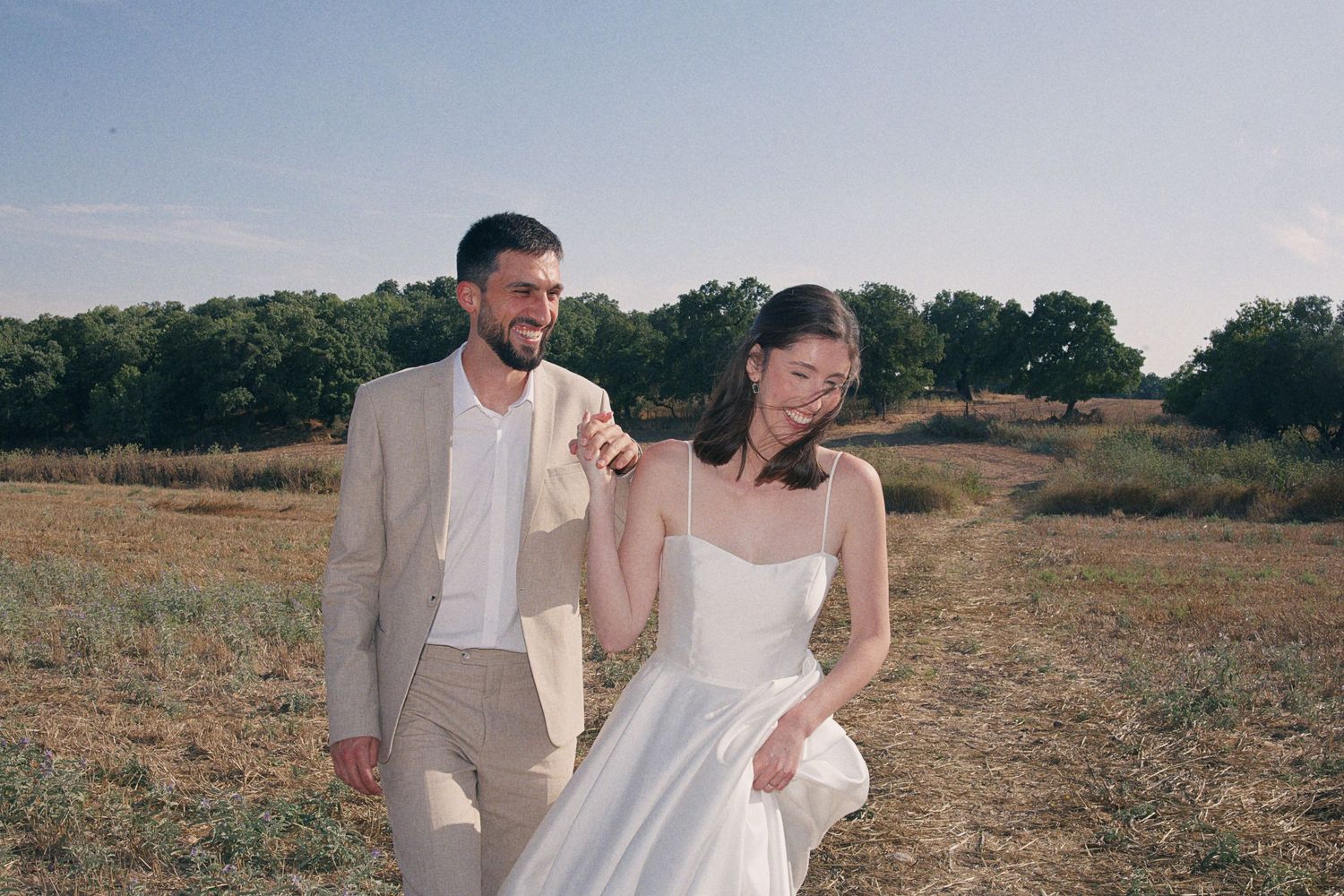 A couple in wedding attire walks through a sunny field, the groom in a tan suit holding the bride's white dress.