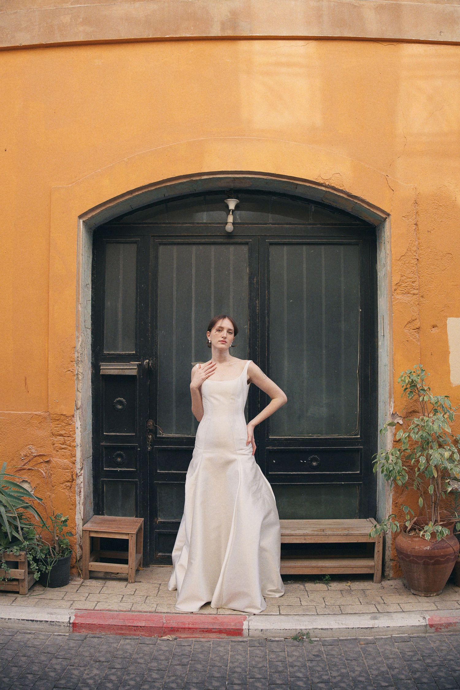 Elegant bride in a sleek white satin gown poses before a vintage green arched door on a cobblestone street.