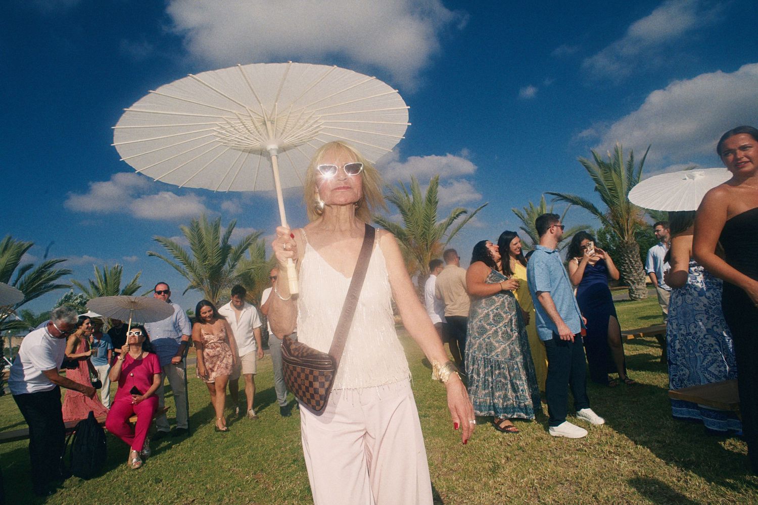 Woman in white outfit holding parasol umbrella at outdoor tropical party with palm trees and blue sky.