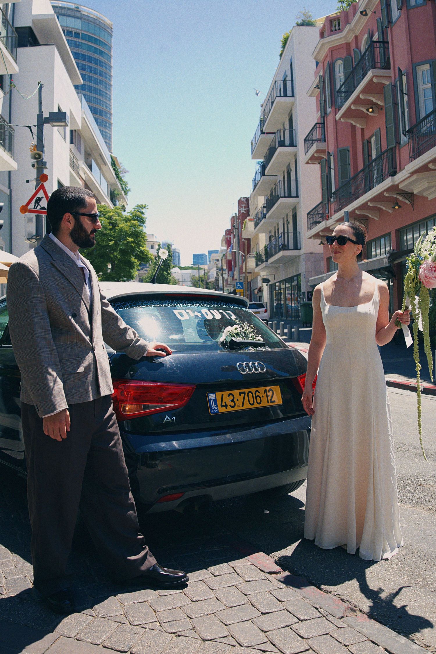 Bride in white gown and groom in gray suit standing beside a decorated Audi car on a sunny city street.