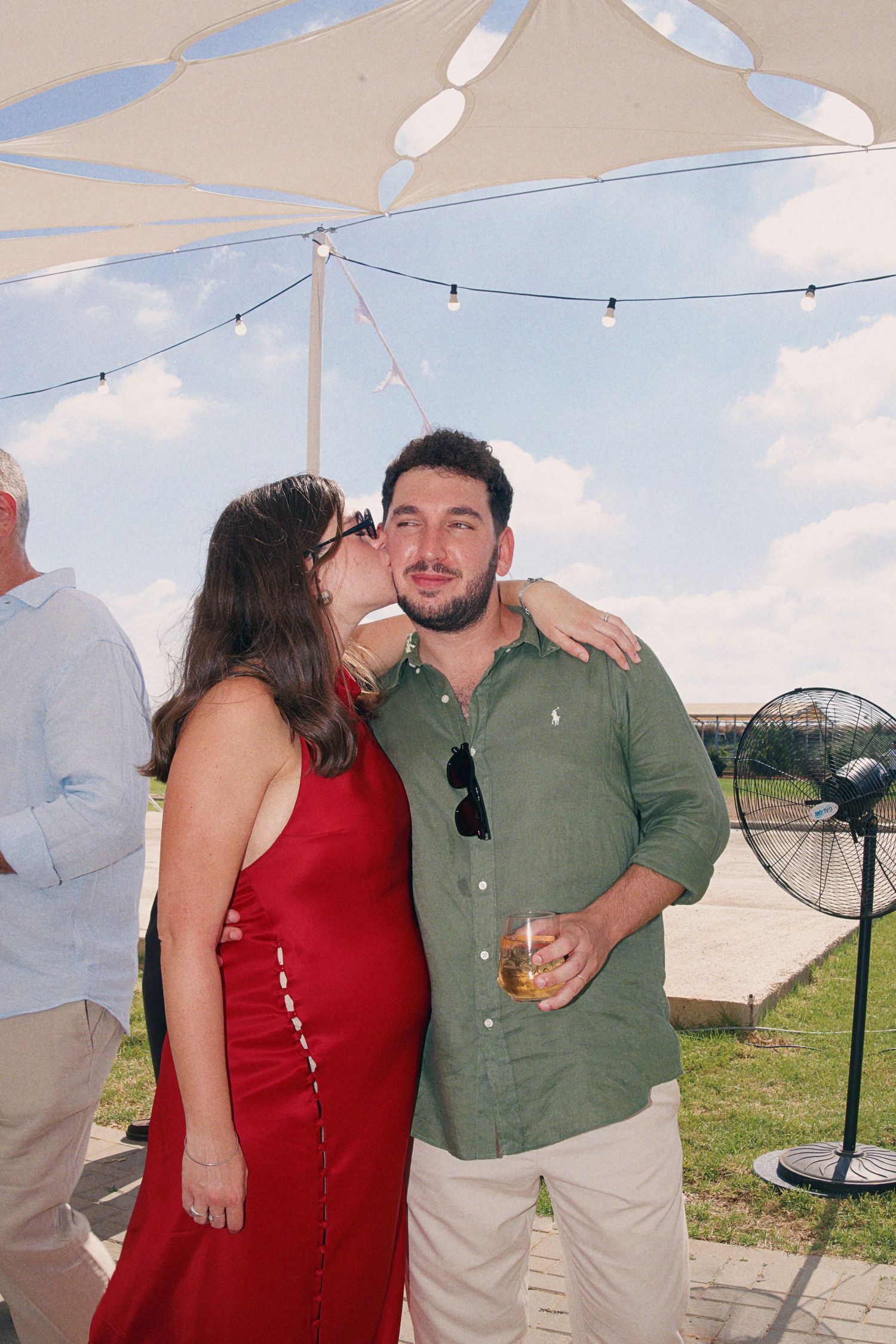 Woman in red dress kisses man in green shirt holding drink at outdoor summer party under white umbrella.