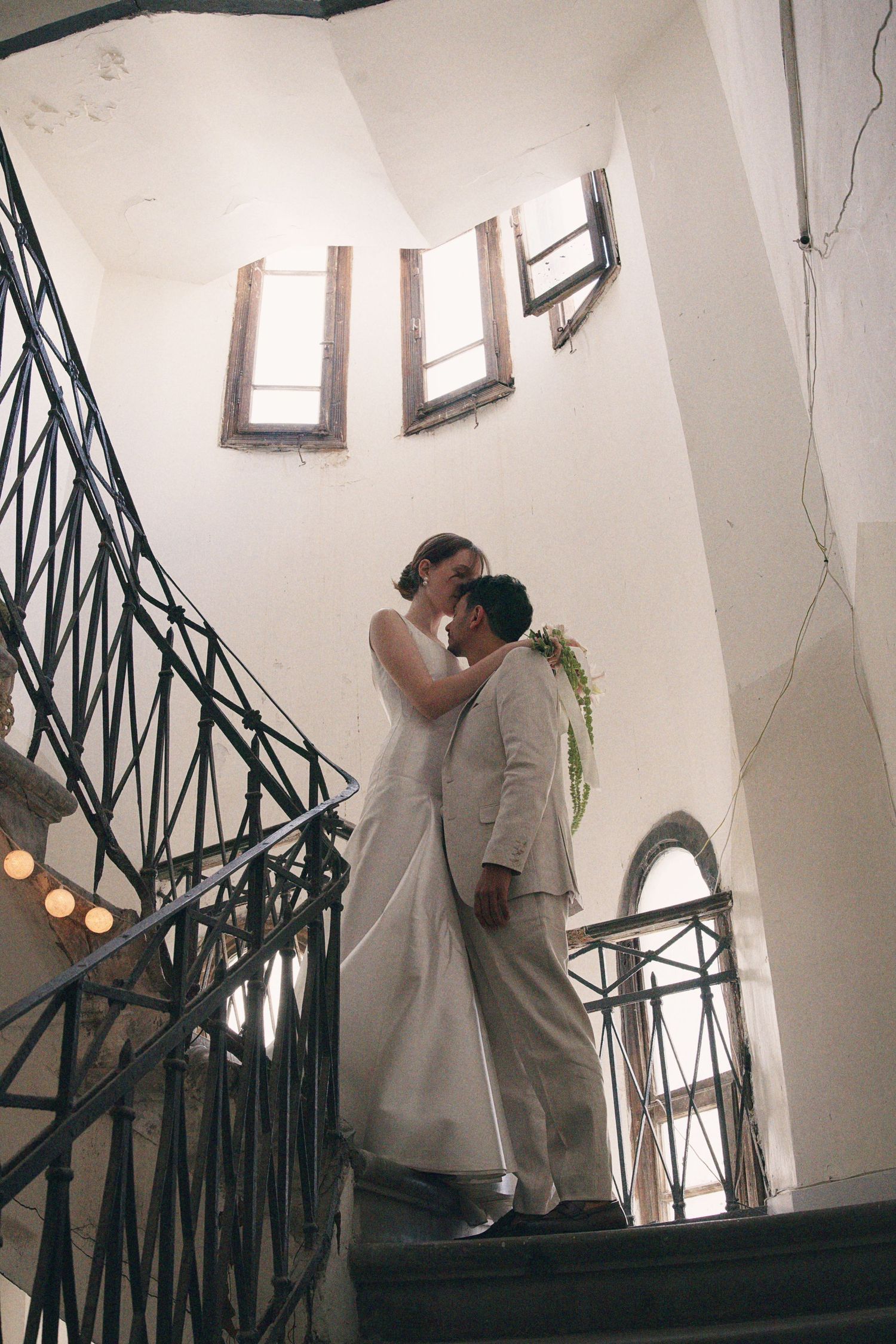 Bride and groom sharing romantic moment on ornate staircase with wrought iron railings.