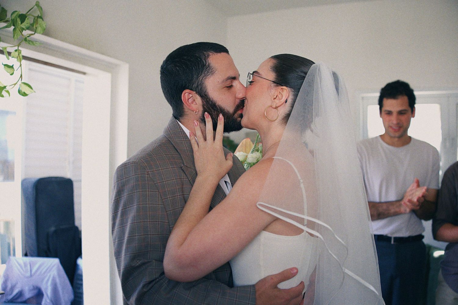 Bride and groom share their first kiss at wedding ceremony while guest applauds in background.