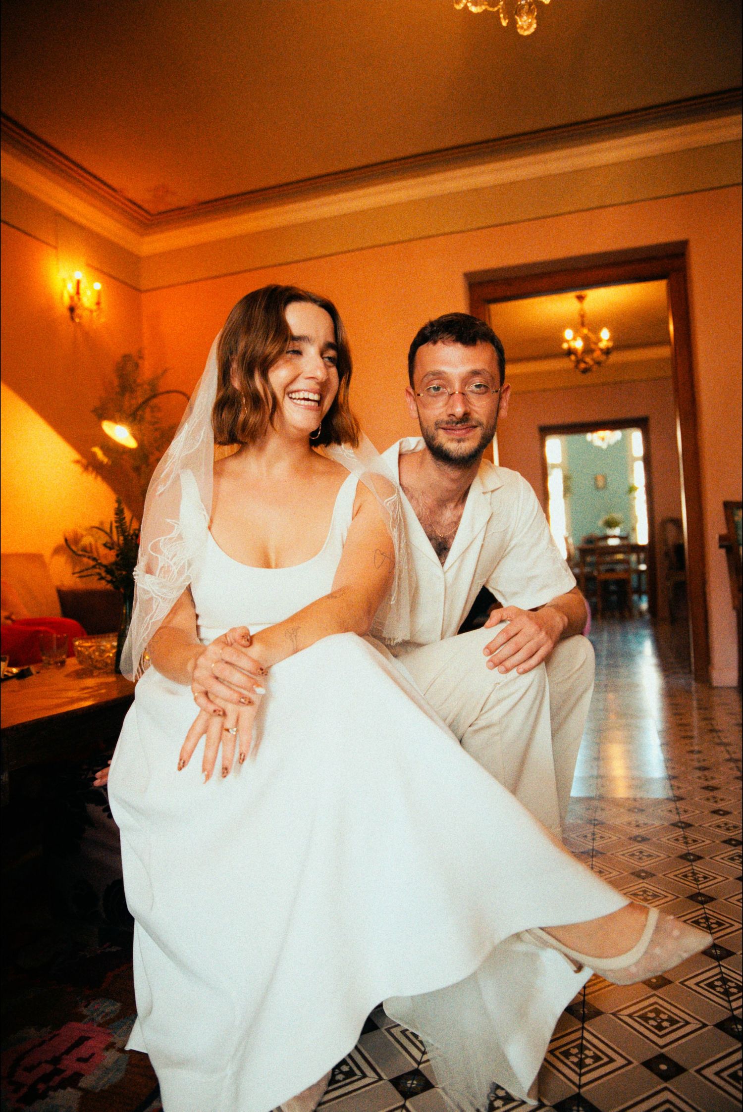 A couple in white attire share a joyful moment sitting on a patterned floor at their wedding celebration.