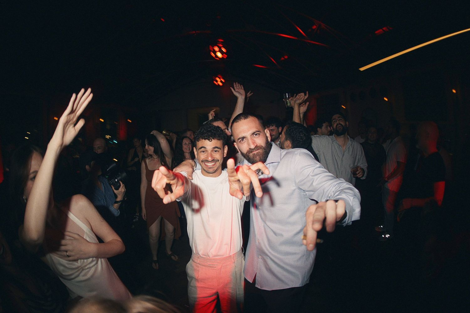 Two men smiling and posing together on a lively nightclub dance floor with crowd and red lights.