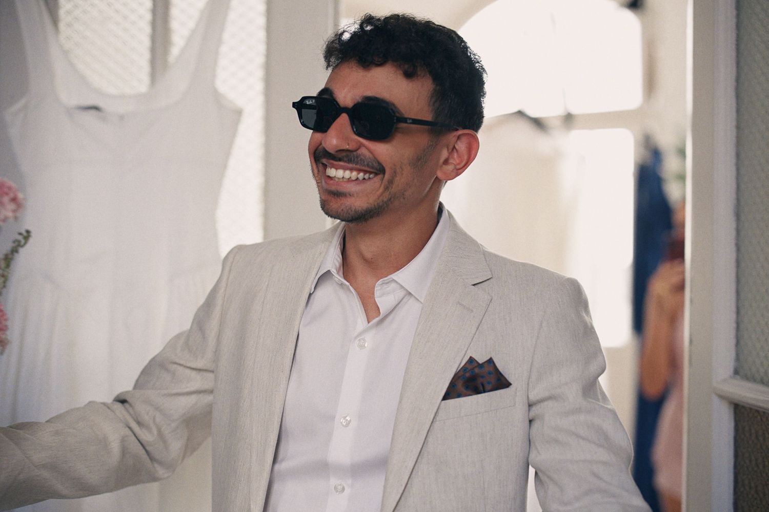 Smiling man wearing black sunglasses and a light beige linen suit with a pocket square at an outdoor event.