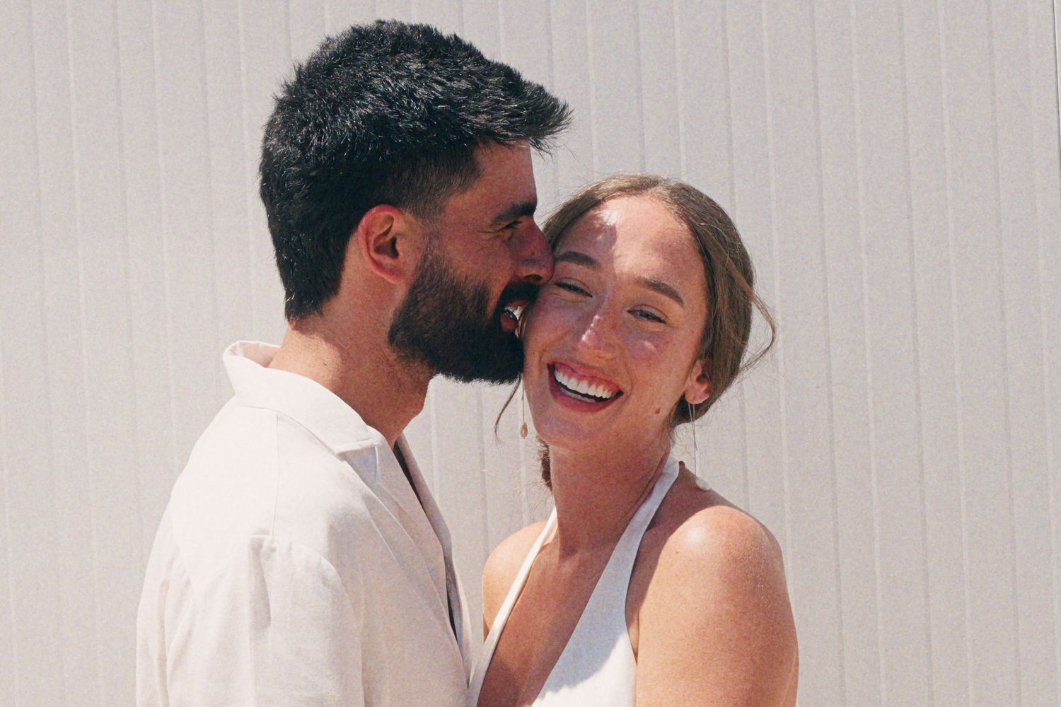 Happy couple sharing a romantic moment, man kissing woman's cheek, both wearing white, smiling outdoors.
