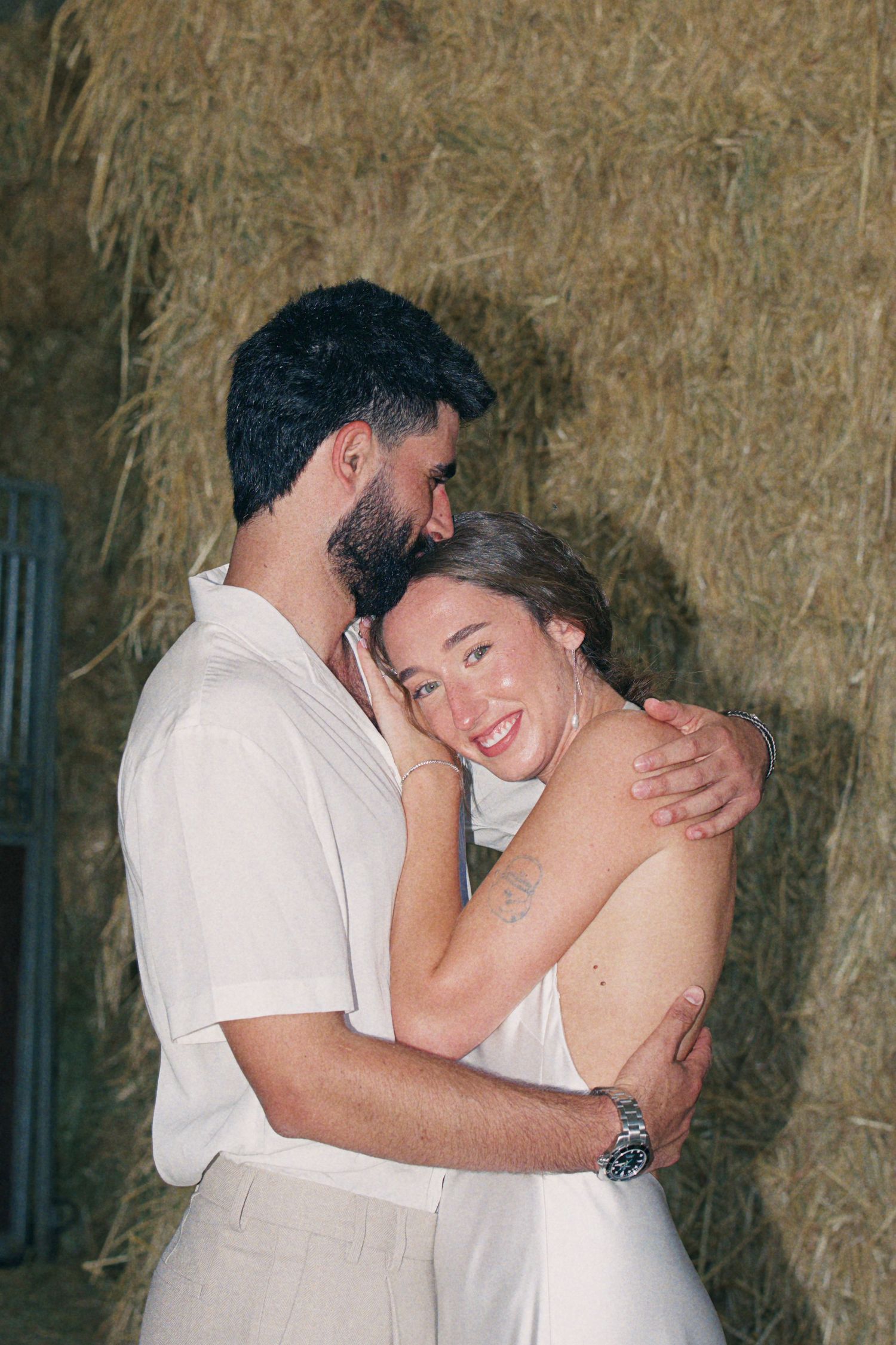 Couple embracing and smiling in front of hay bales, wearing white outfits at a rustic event.